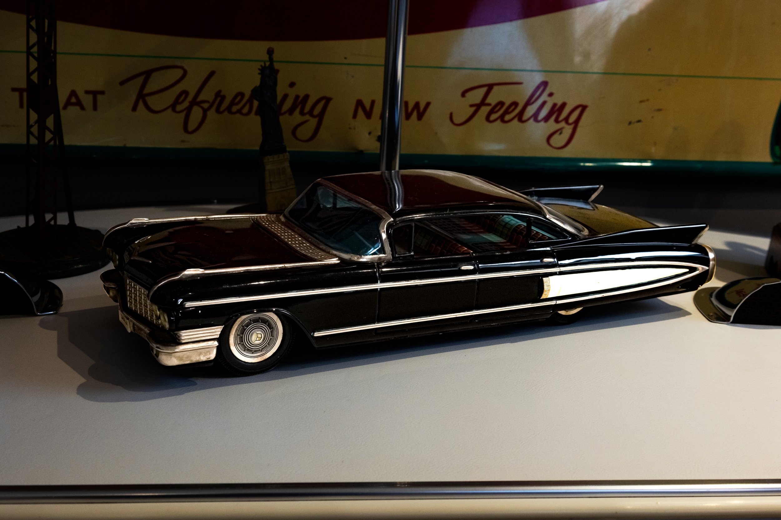 Model van een klassieke zwarte Cadillac auto met een dak van bruin leer, geplaatst op een witte displayplaat, met een achtergrond met tekst die gedeeltelijk zichtbaar is.