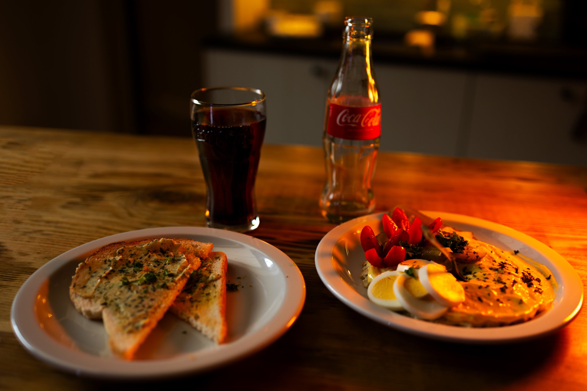Verlichte tafel met twee borden eten, een glas cola en een fles Coca-Cola. Eén bord met gesneden brood en kruiden, ander met een salade met tomaat, hardgekookte eitjes en garnalen.