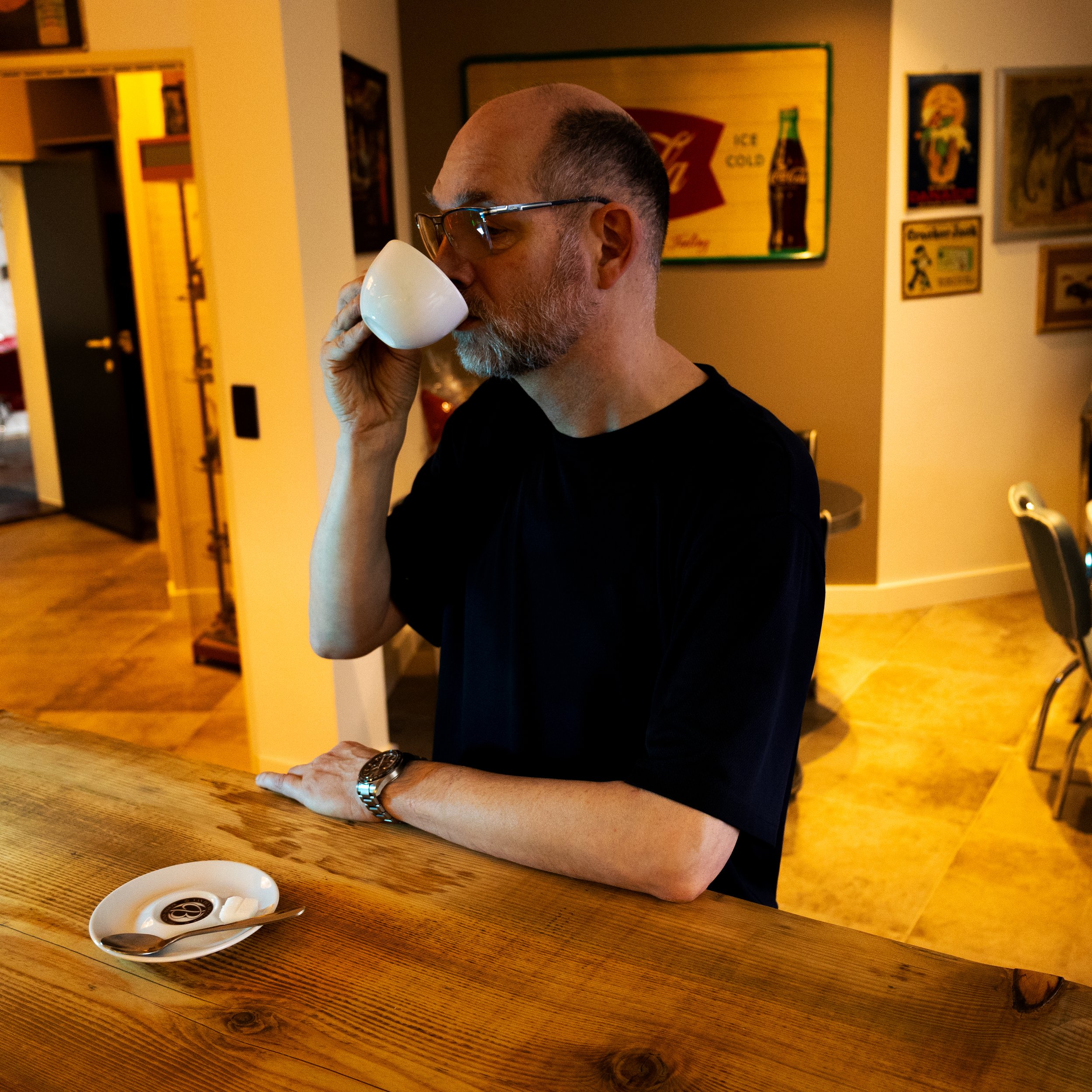 Man met grijs haar en een baard drinkt uit een witte kop in een cafe of restaurant, zittend aan een houten bar.