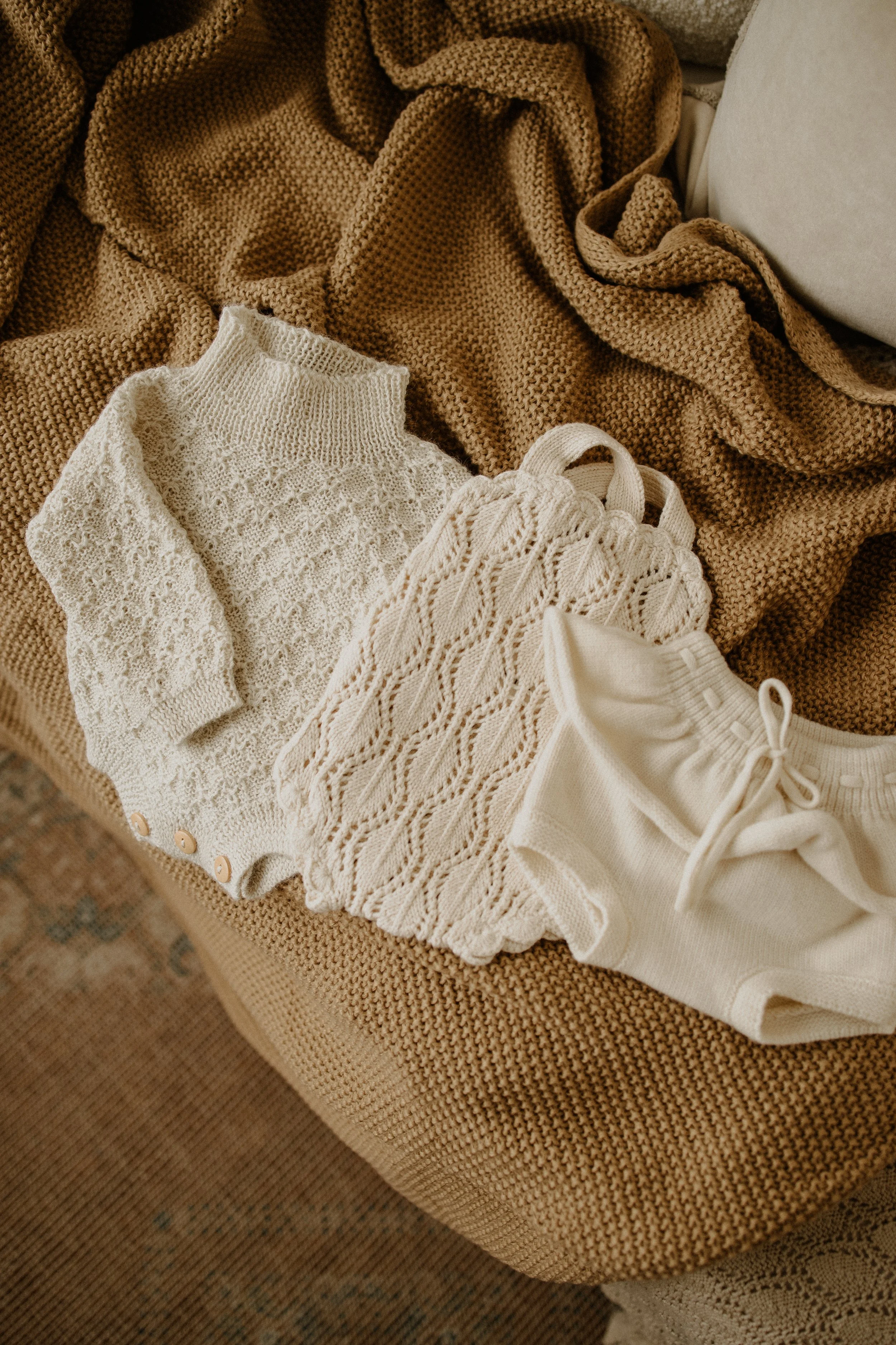 A collection of cream-colored baby clothes including a knitted sweater, a cable-knit romper, and a pair of shorts, laid out on a textured beige blanket.