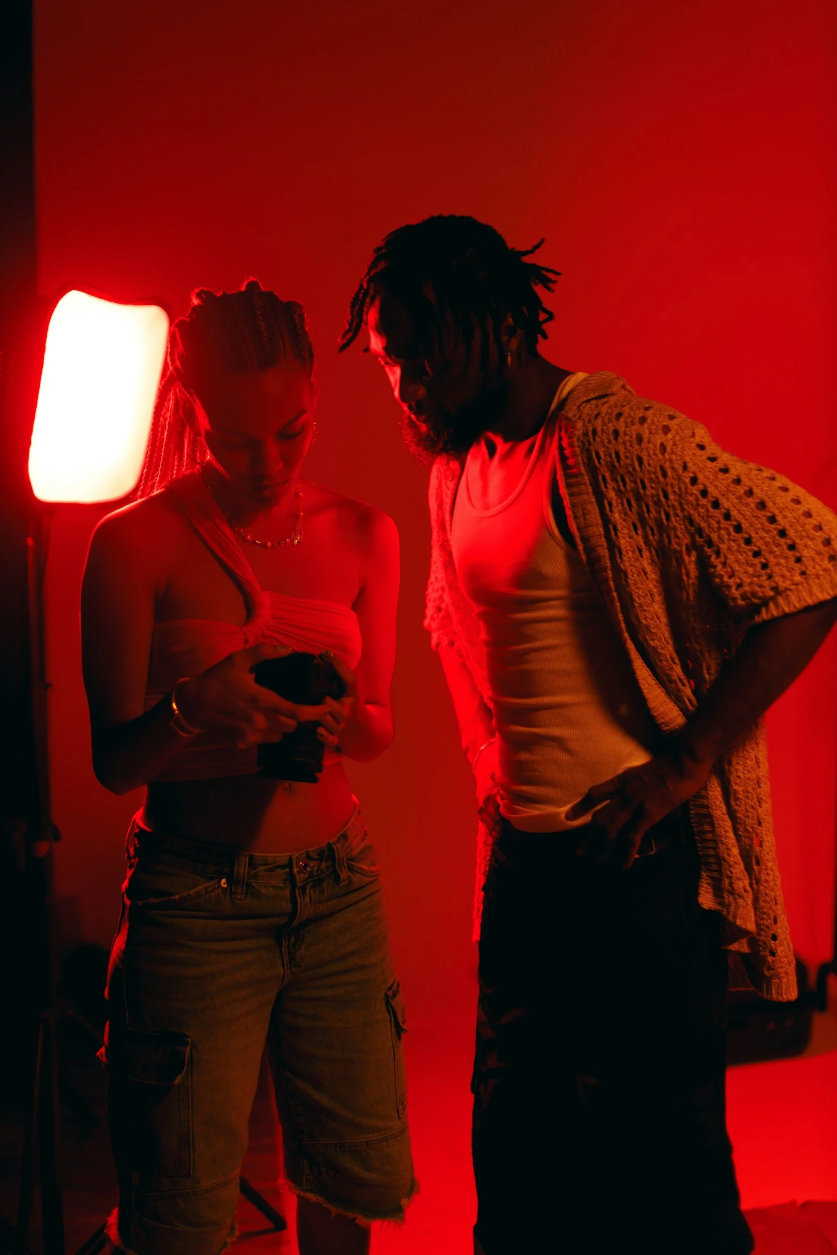 Two people are standing close together in a dimly lit room with red lighting, looking down at an object they are holding, possibly a phone. One person has braided hair and is wearing a strapless top and cargo shorts, and the other has dreadlocks and is wearing a tank top with a crochet cardigan.