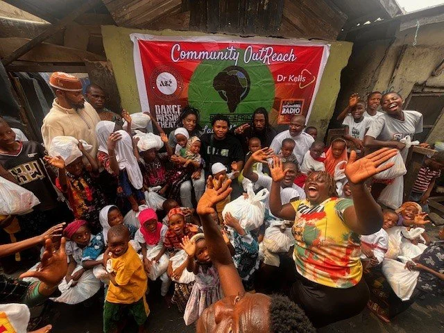 A large group of children and adults gathered indoors, smiling and waving, in front of a red banner that reads 'Community Outreach Dr. Kells.' The group appears to be at a community event or outreach program.