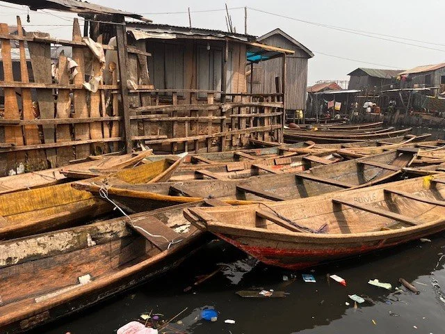 Several wooden boats docked along a muddy waterway with a makeshift wooden structure in the background.