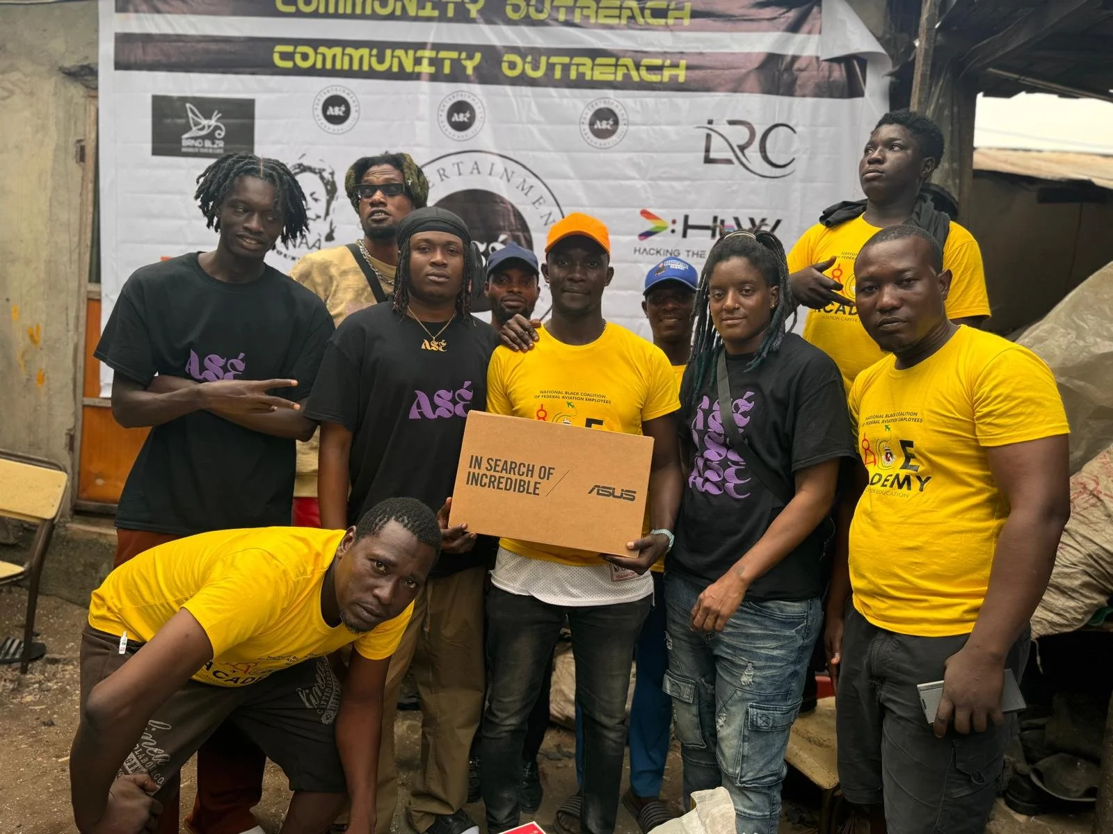 Group of young people posing together at a community outreach event, holding a cardboard box with the ASUS logo. They are in front of a banner with multiple logos and text, some wearing black and yellow t-shirts with event branding.