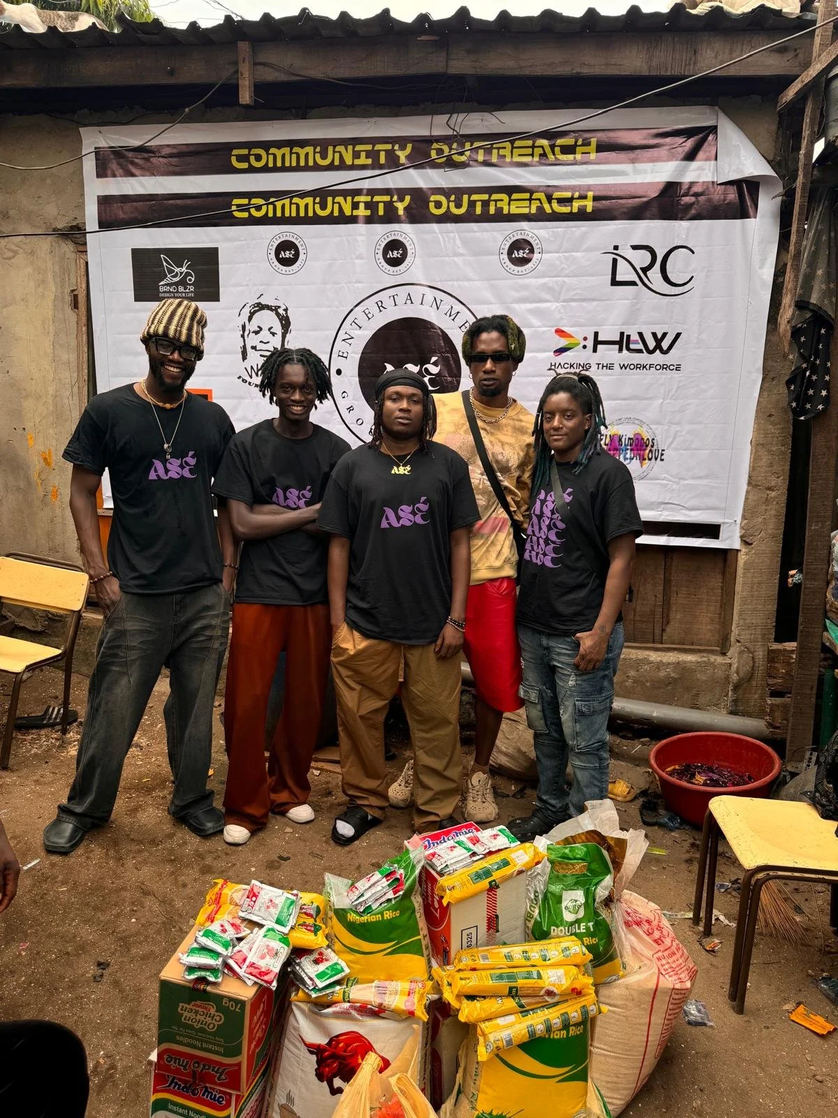 A group of five people standing in front of a donation banner with various logos. In front of them, there are bags of rice and food supplies on the ground. The setting appears to be an outdoor, rustic area with chairs, a bowl with food, and a makeshift structure.
