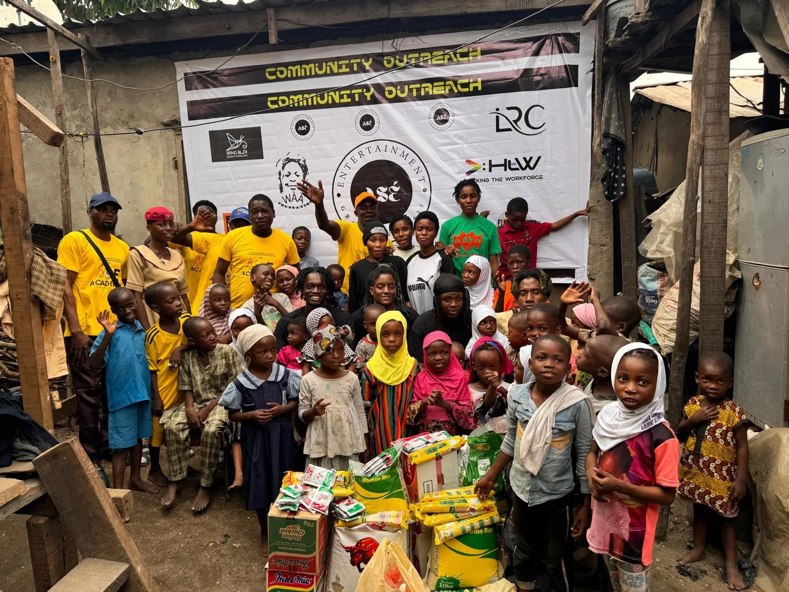 Group of children and adults gathered in front of a banner with community outreach logos, with food supplies on the ground in front of them, in a modest setting with makeshift structures.