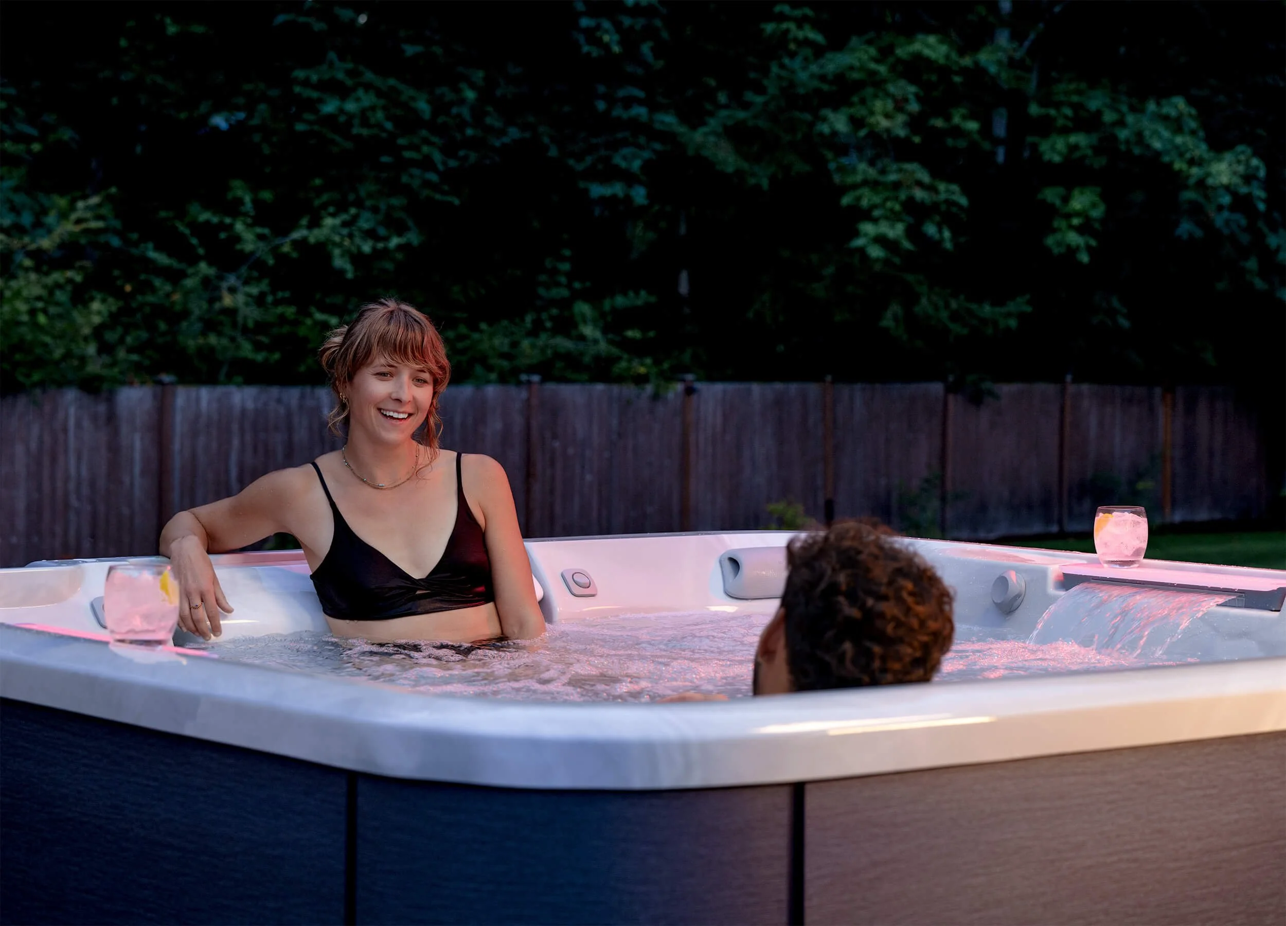 Woman in black bathing suit lounging with man in Jacuzzi® J508L Hot Tub in a residential backyard setting with trees and fence in background.