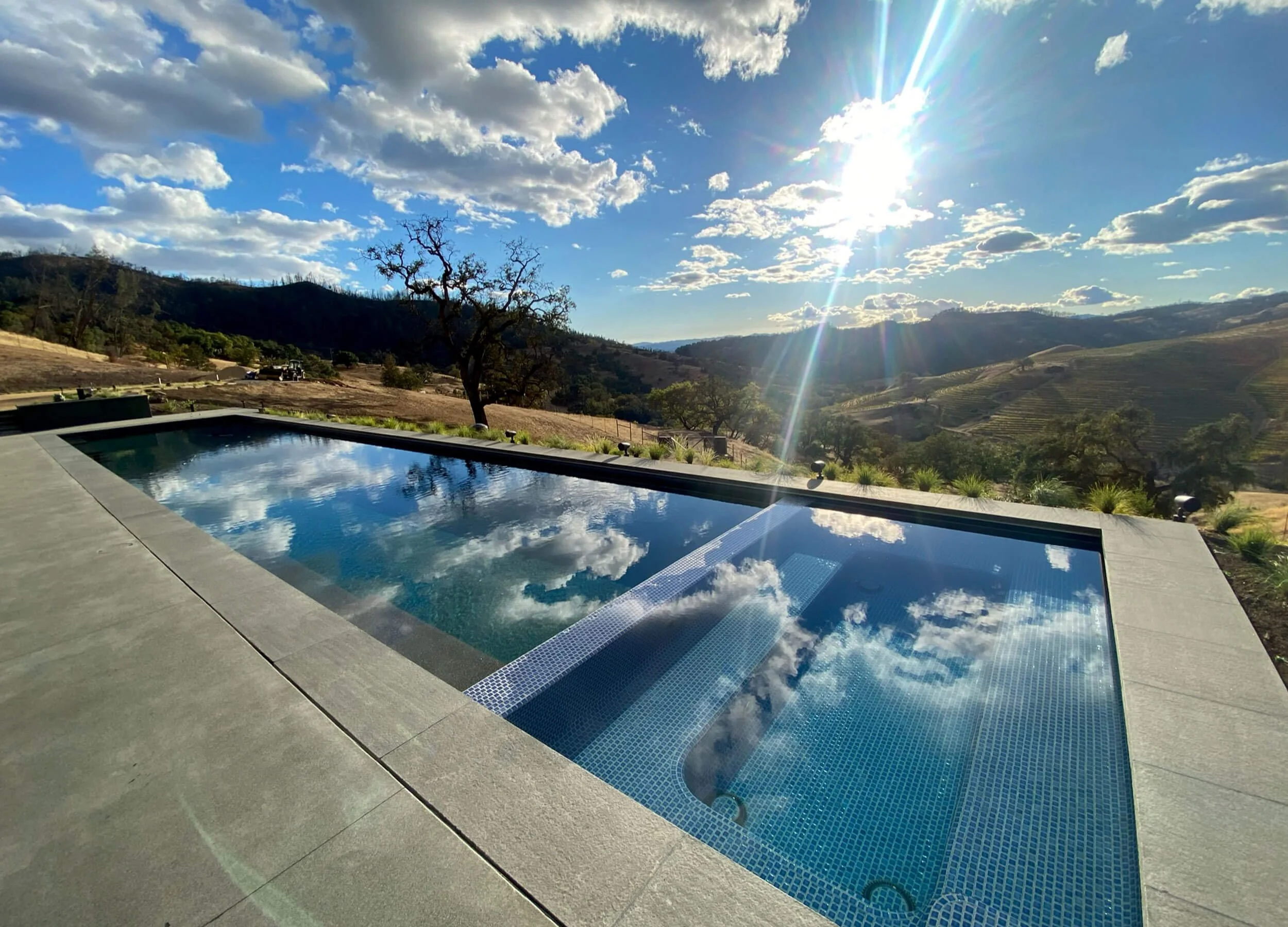Image of sun and clouds reflecting off the surface of a custom pool build by Johnson Pool & Spa - pastoral rolling hills, blue sky with white clouds, grassy hillsides with trees in view.