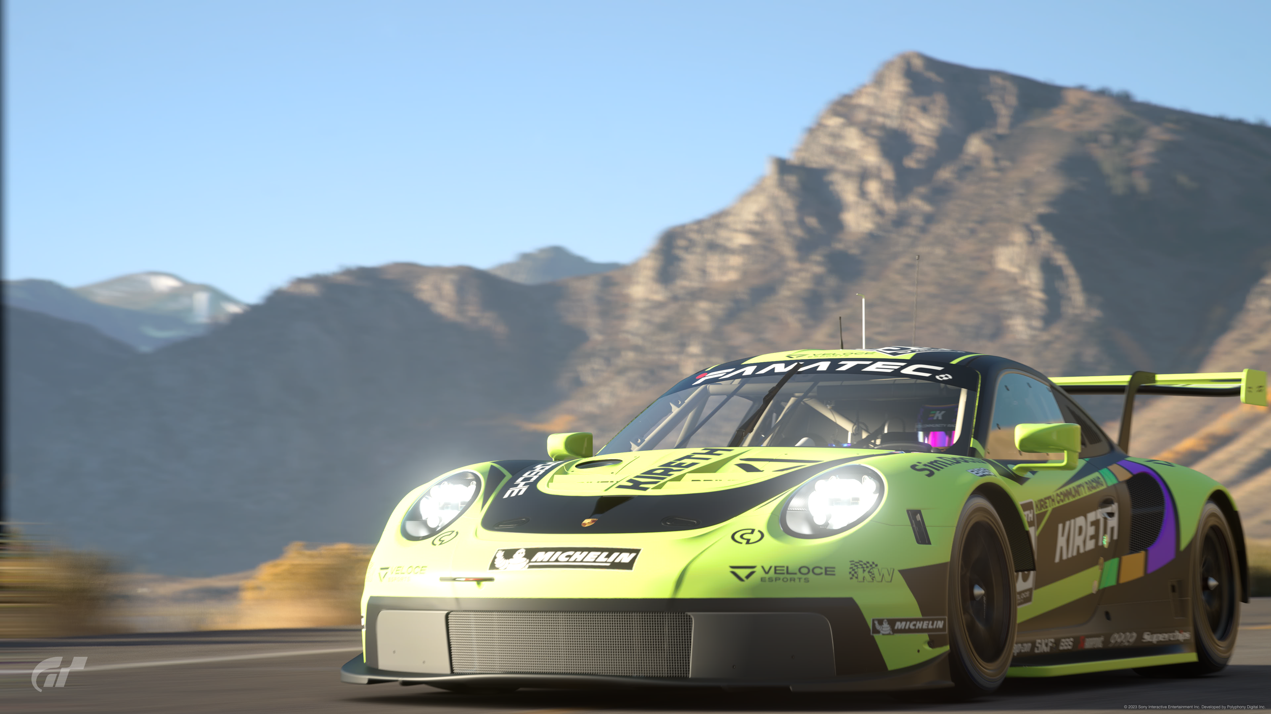 Green and black race car speeding on a mountain road with rocky mountain landscape in the background.
