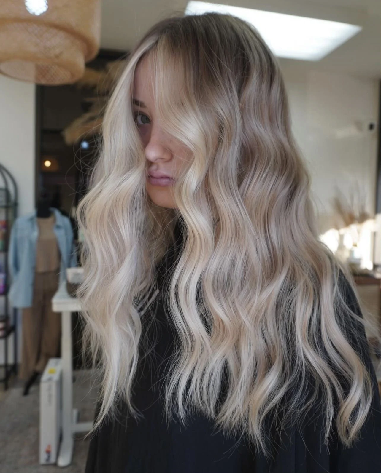 A woman with long, wavy blonde hair, partially covering her face, standing in a salon or similar setting.