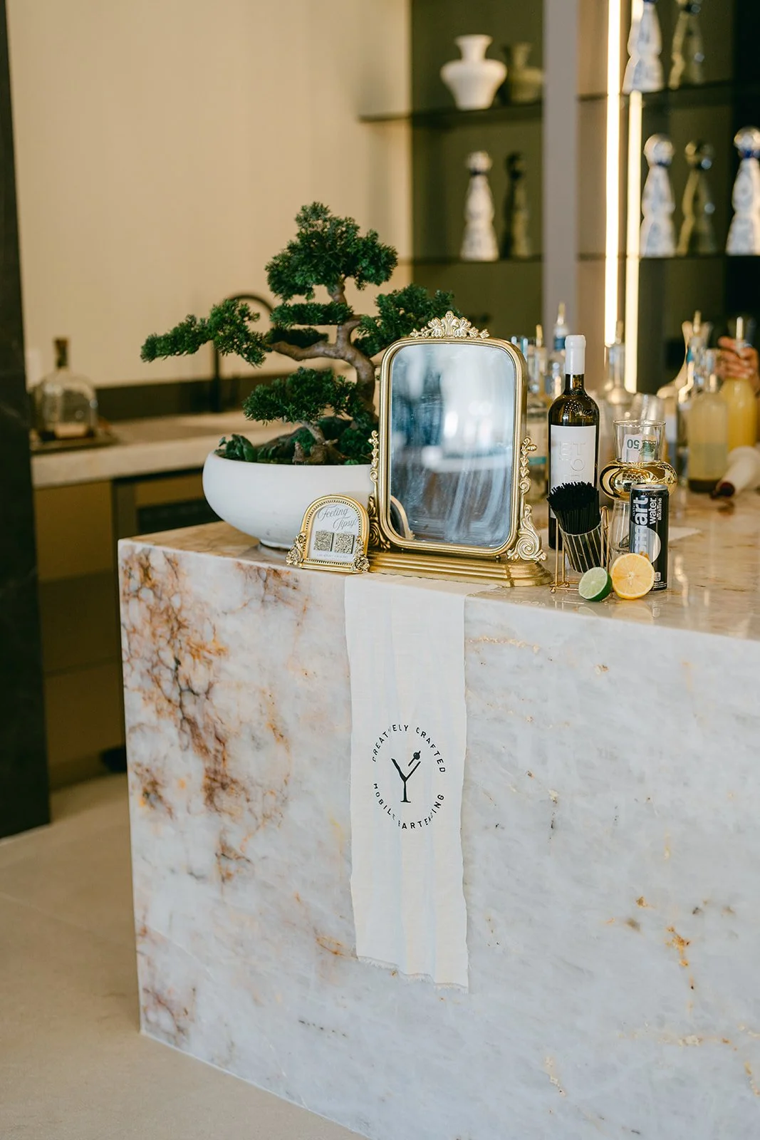 A marble bar counter with a bonsai tree in a white pot, a small ornate mirror, and various bottles and bar tools, including lime and lemon slices, a cocktail shaker, and stirrers.