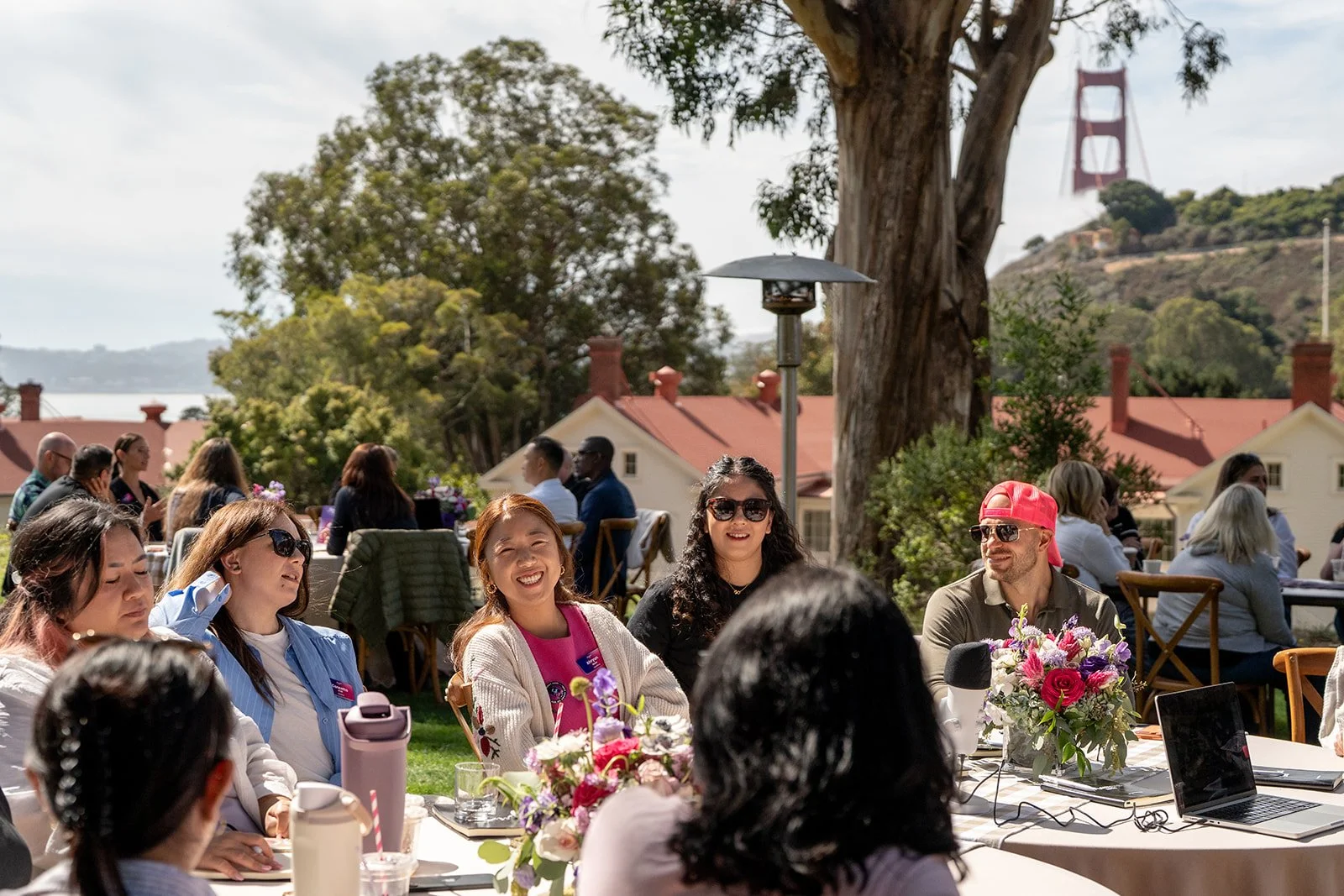 People gathered outdoors at a social event with a view of the Golden Gate Bridge in the background. The group is sitting around tables with flowers, smiling and chatting, with some wearing sunglasses and casual clothing.