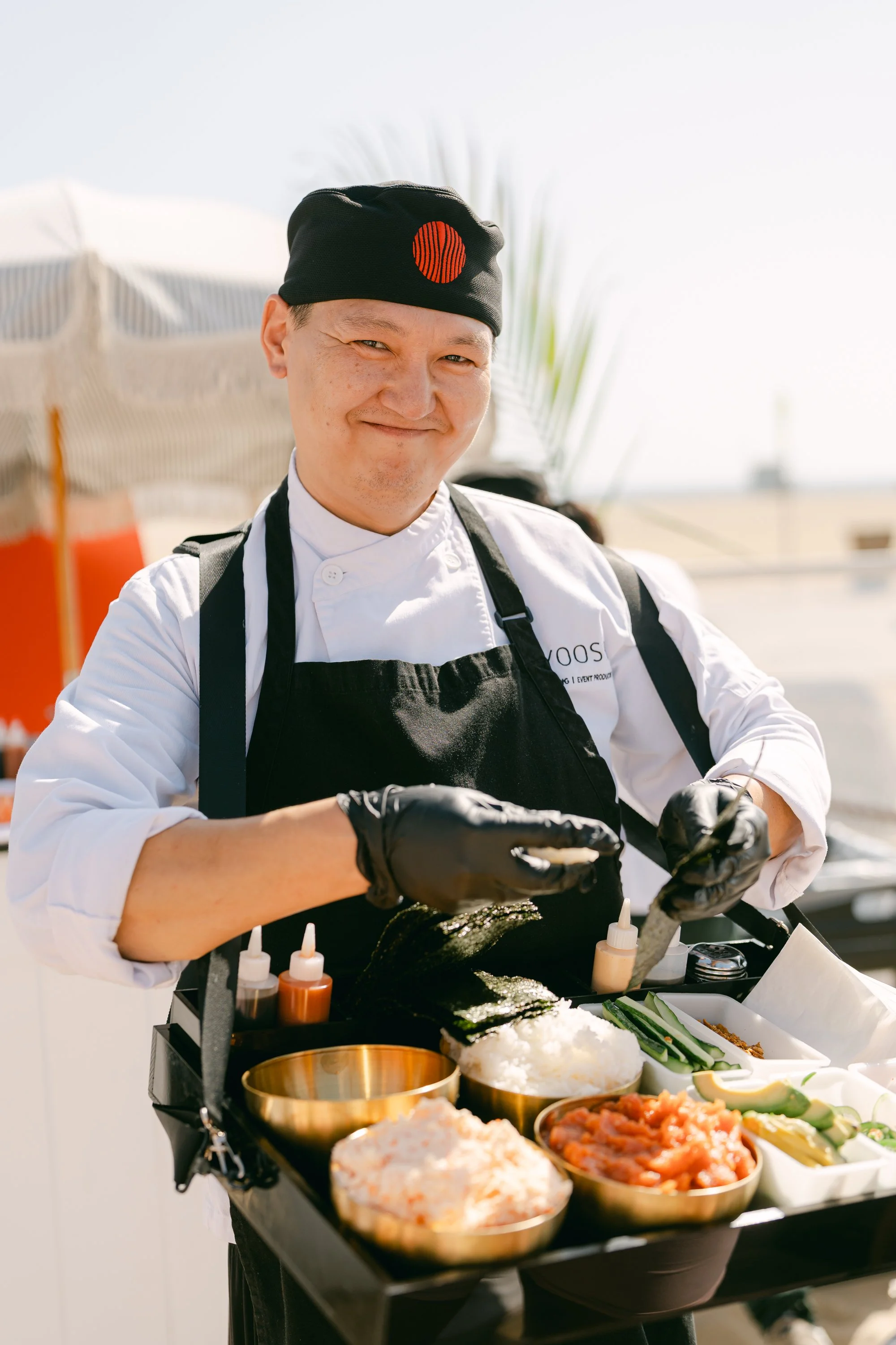 A chef wearing a black hat with a red circle design and a white chef's coat with a name tag is preparing food outdoors. The table displays various ingredients including rice, vegetables, and sauces, with a blurred background of umbrellas and a beach 