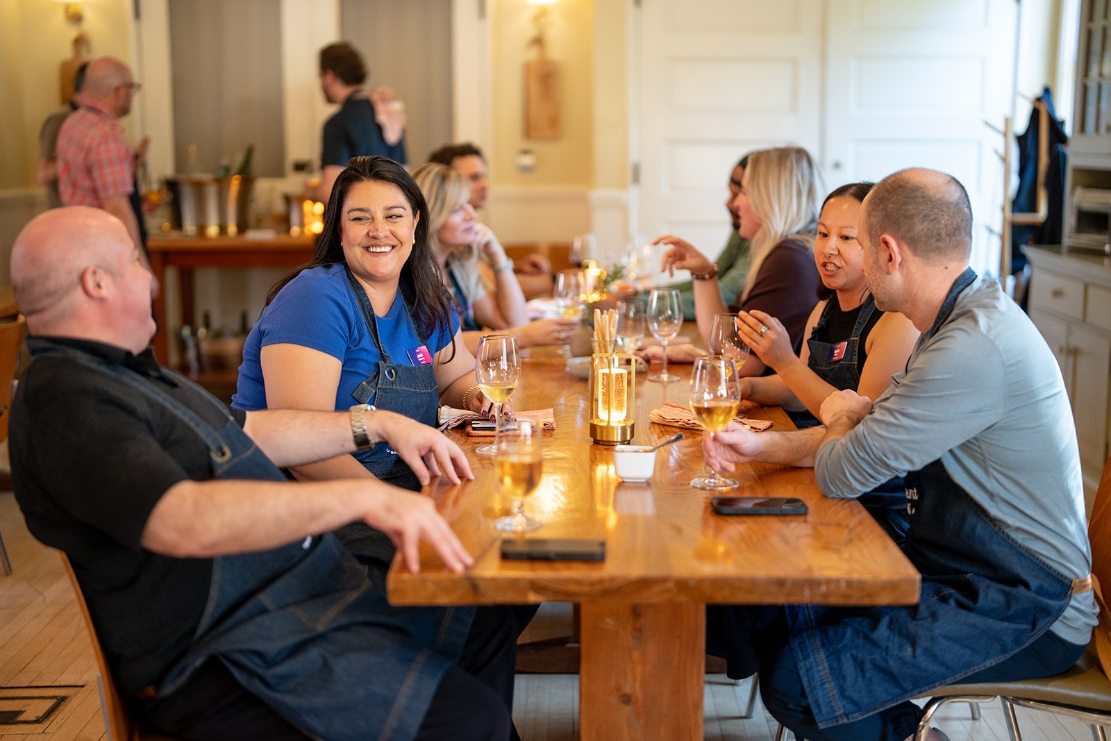 Group of people sitting around a wooden dining table, enjoying drinks and conversation in a warmly lit room.