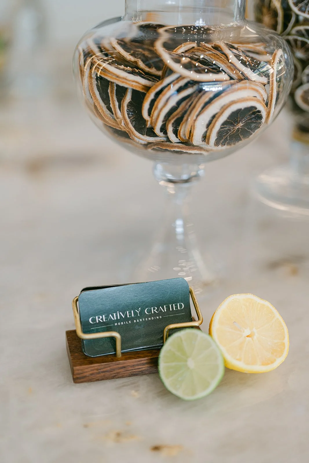 A glass jar filled with dried lemon slices, a small wooden block with a business card reading 'Creatively Crafted Mobile Bartending', and halves of lemon and lime on a table.