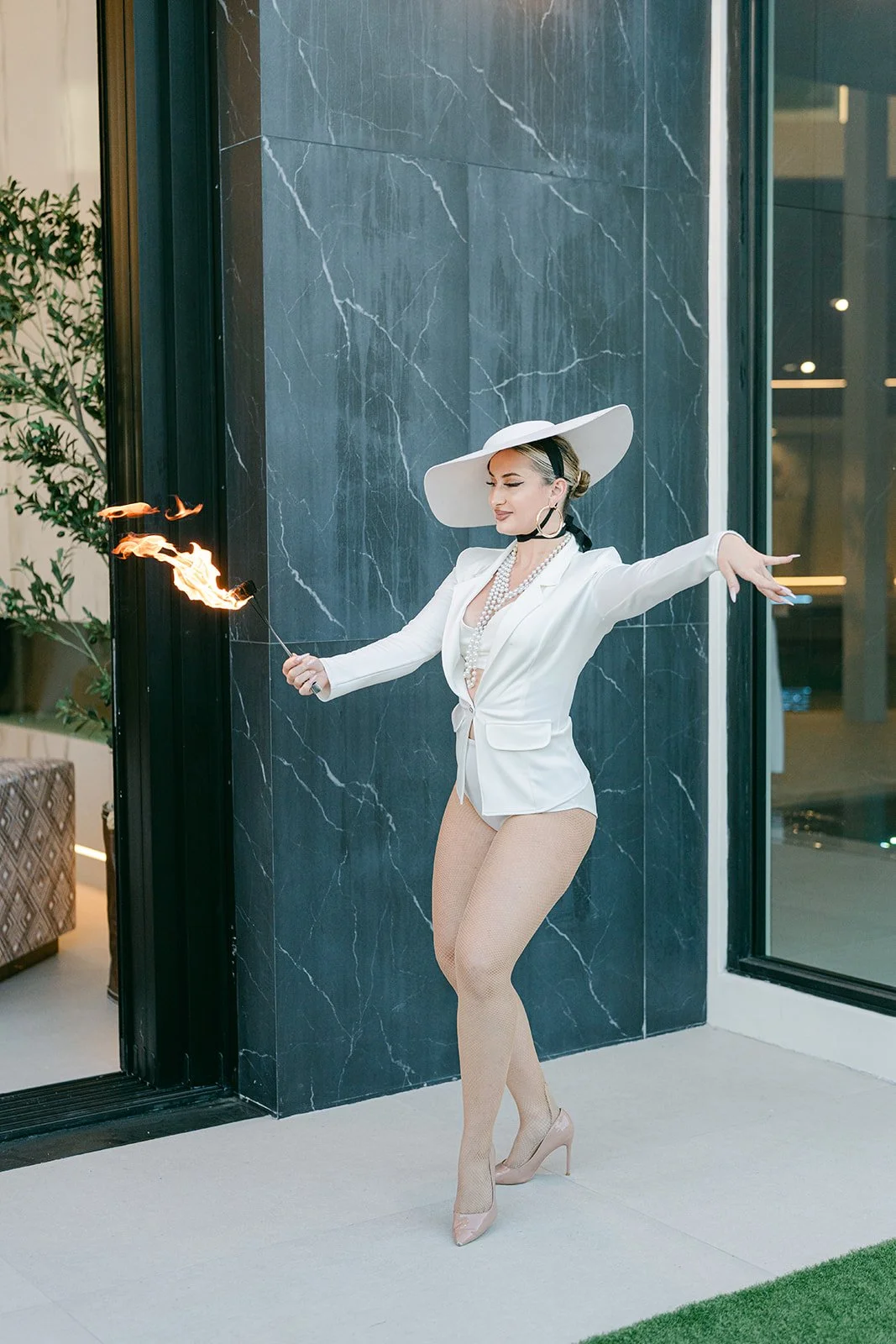 Woman in white blazer, beige fishnet stockings, and high heels stands outside a building holding a flaming torch, wearing a large white hat and jewelry, performing in front of a black marble wall.