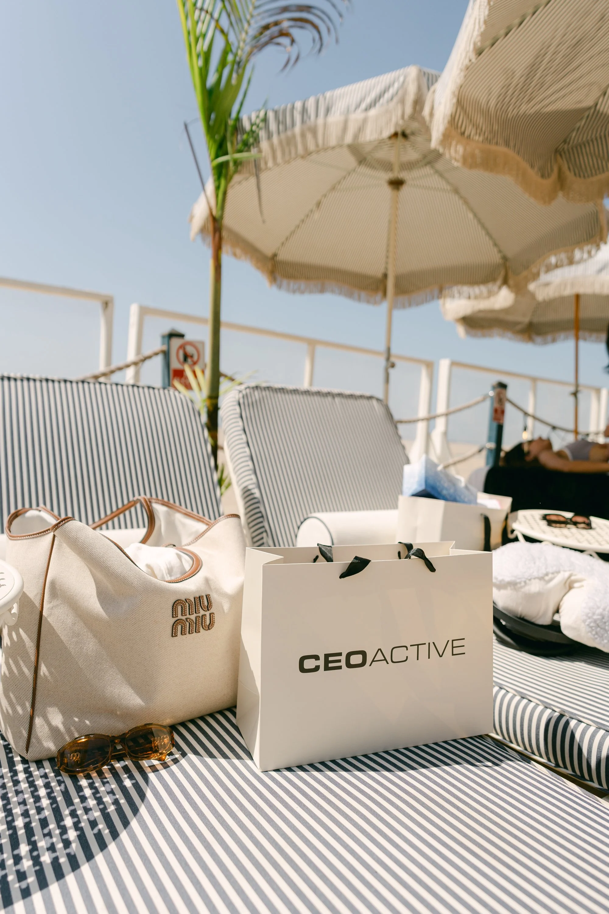 Beach lounge area with striped umbrellas, chairs, and a table with shopping bags, sunglasses, and a CE0ACTIVE shopping bag, under a clear blue sky.