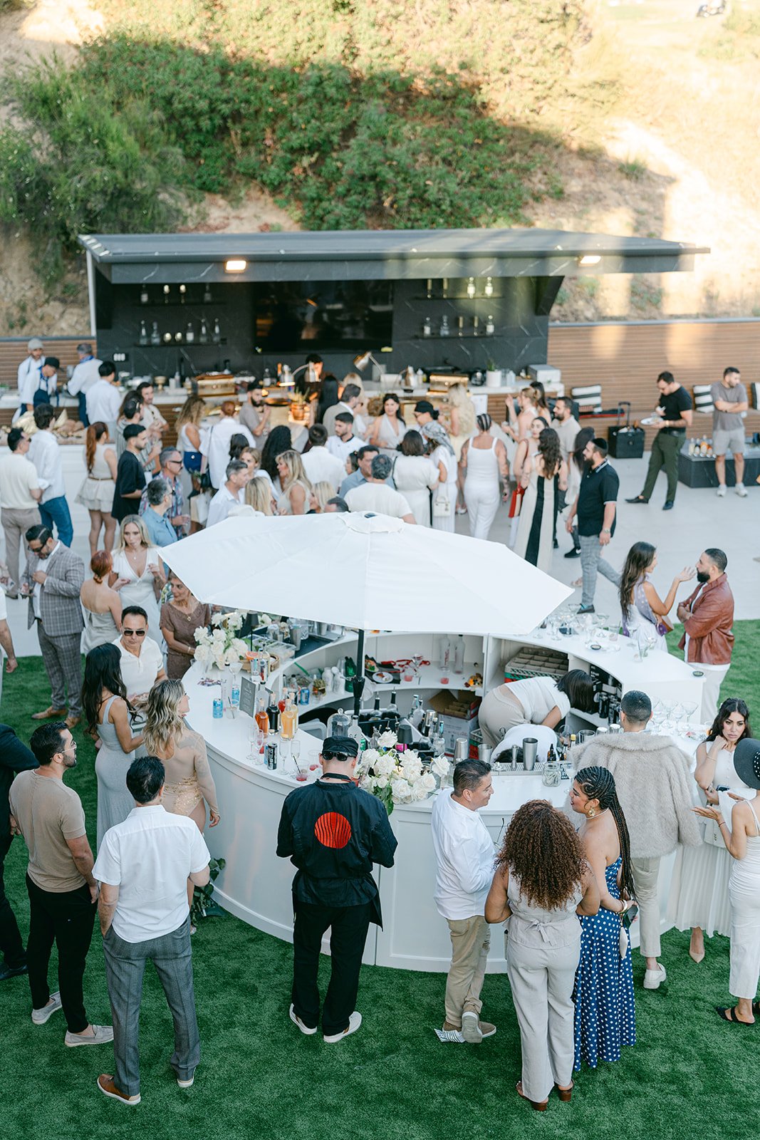 People attending an outdoor event with a bar and food area, some socializing and others waiting in line, set against a hillside backdrop.