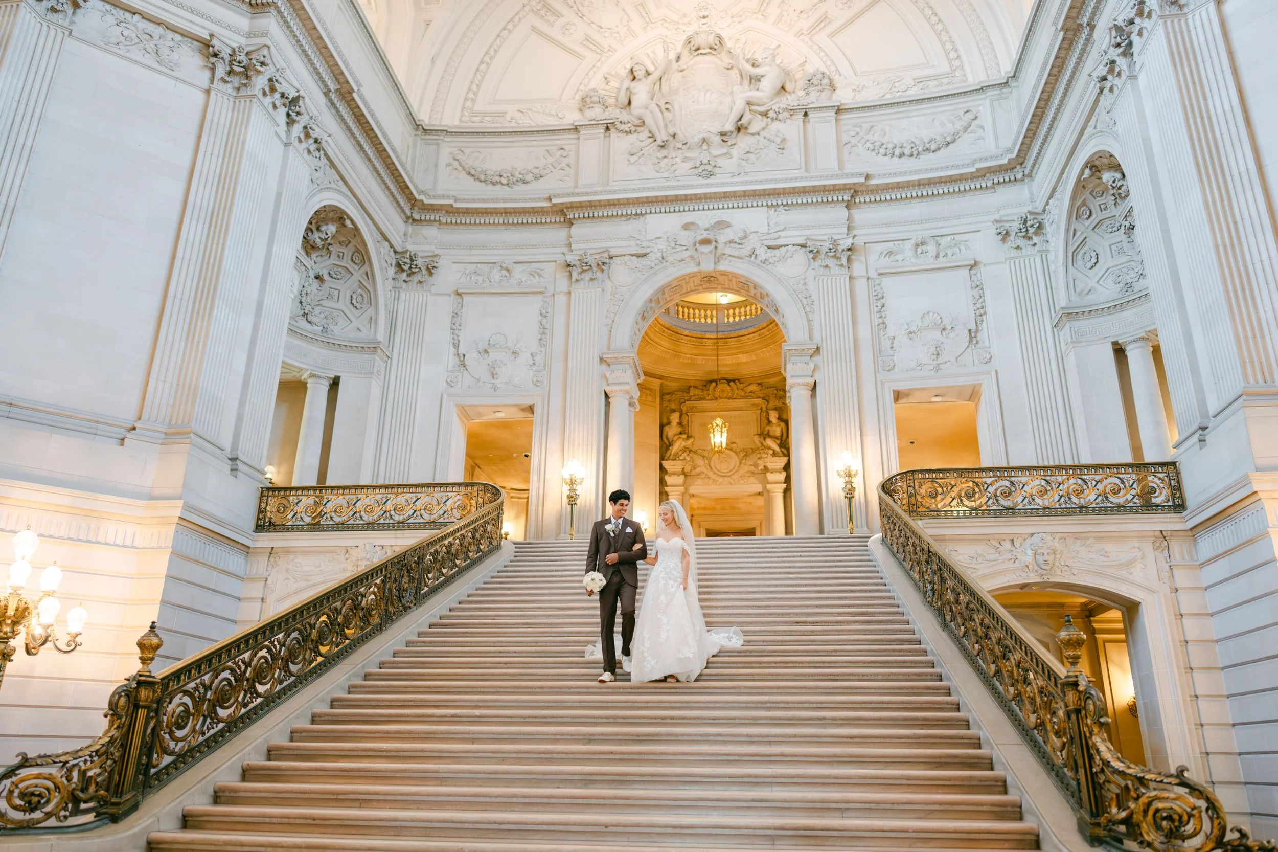 A wedding couple standing on grand staircase inside a lavish, ornate building with high ceilings and intricate architectural details.