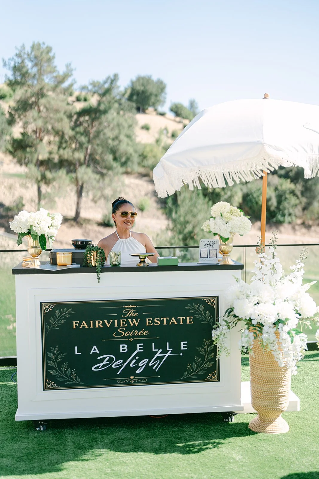 A woman smiling at a decorated outdoor bar with white flowers, a large white umbrella, and a black sign with gold and white lettering reading "The Fairview Estate Soirée La Belle Delight." The background features green trees and a light blue sky.