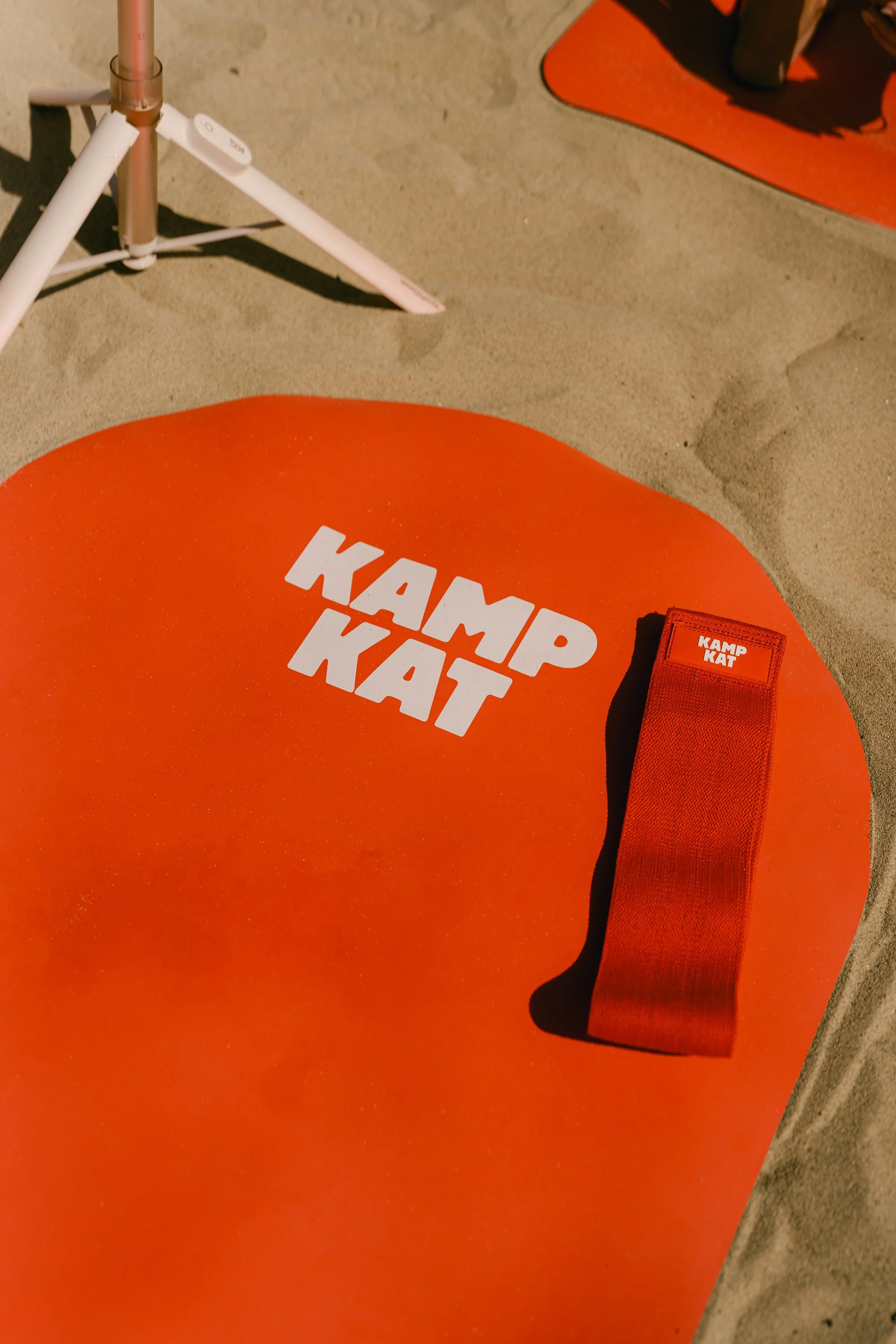 A bright orange circular object with the words 'KAMP KAT' written in white, placed on a sandy surface. Next to it, there is a red strap with a label that says 'KAMP KAT'. In the background, part of an orange mat and a person kneeling are visible.