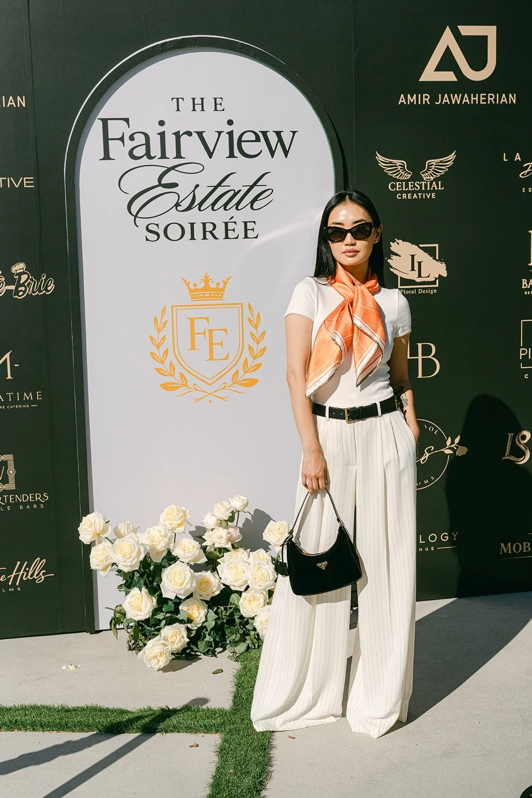 A woman with long dark hair wearing black sunglasses, a white t-shirt, white pinstripe wide-leg pants, a black belt, and an orange scarf around her neck stands beside a white sign that reads 'The Fairview Estate Soiree.' She holds a small black handb