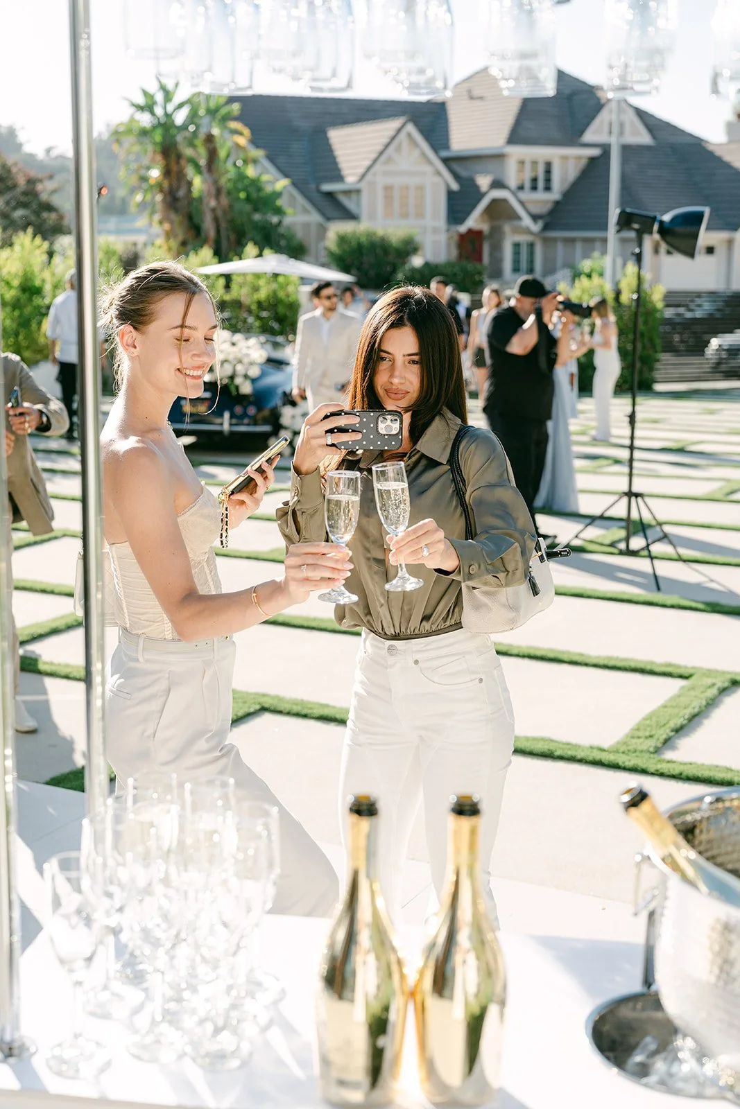 Two women celebrating with champagne at an outdoor event, with one taking a photo and the other smiling, in front of a house with guests and photographers in the background.