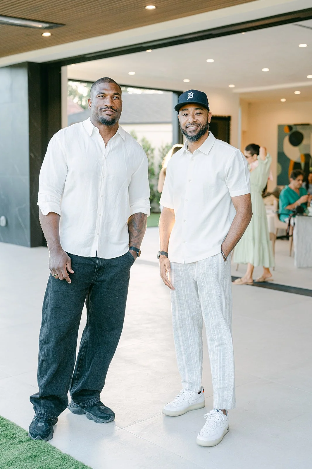 Two men standing and posing for a photo at an indoor event, with other people in the background. One is wearing a white shirt and black pants, and the other is wearing a white shirt, striped pants, and a black baseball cap.