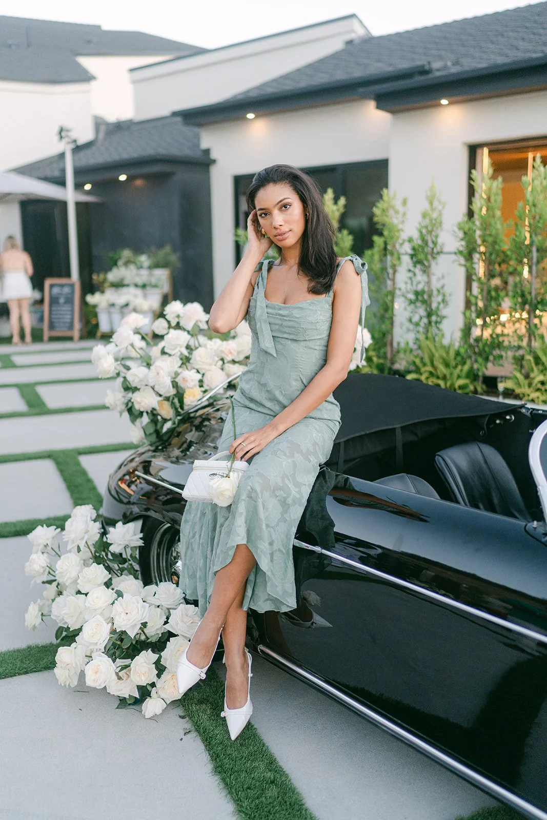 A woman in a light green dress, sitting on a black convertible car with a large bouquet of white roses, outdoors in front of a modern house.