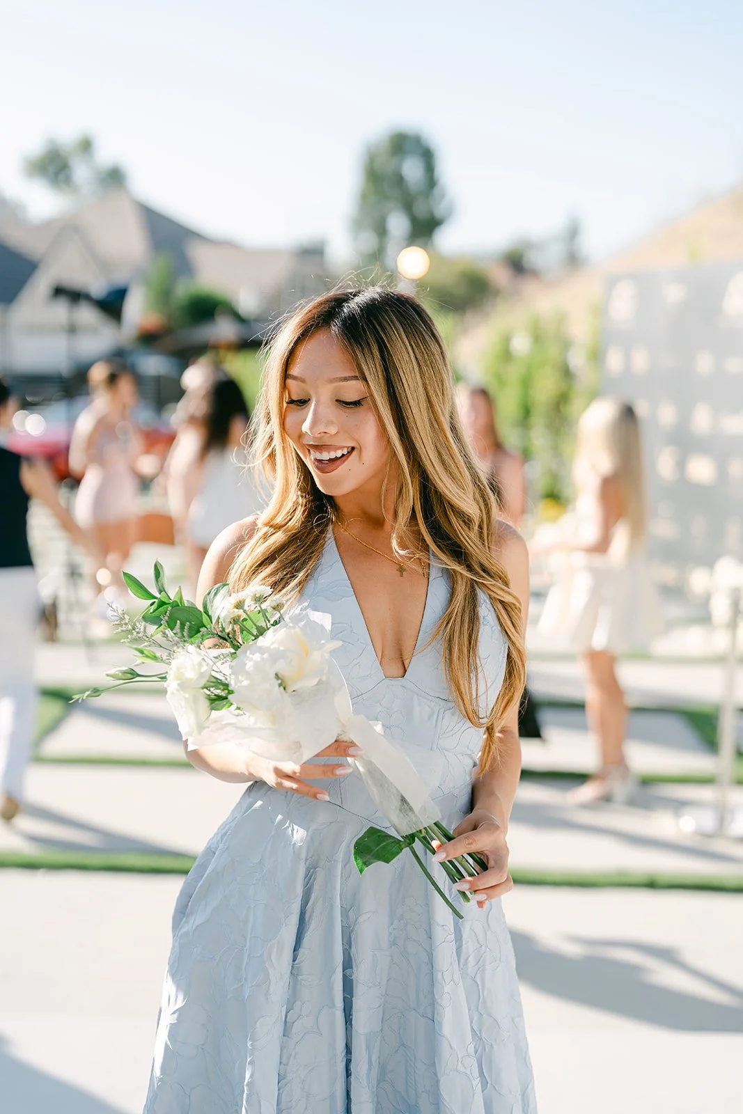 A woman in a light blue dress holding a bouquet of white flowers at an outdoor event with other women in the background.