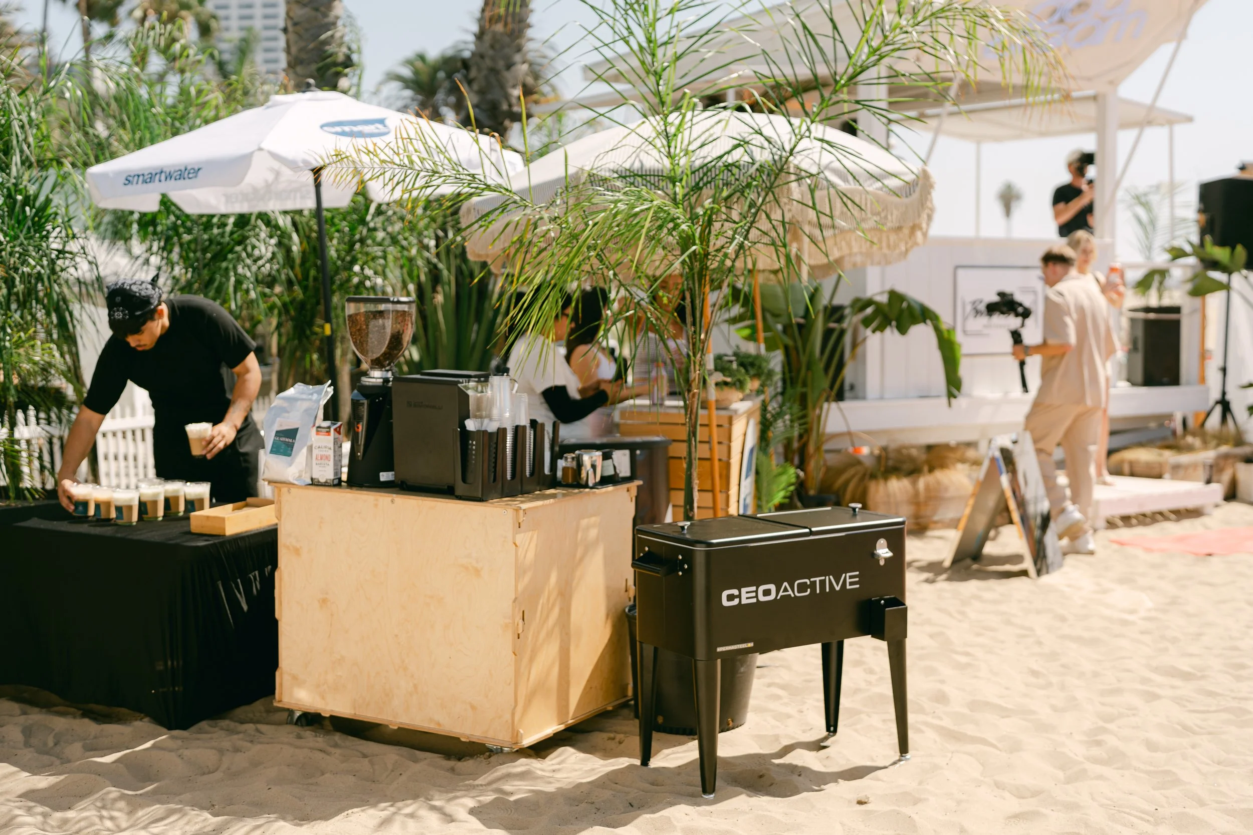 Beachside coffee stand with baristas preparing drinks, surrounded by palm trees, with a person filming in the background under an umbrella.