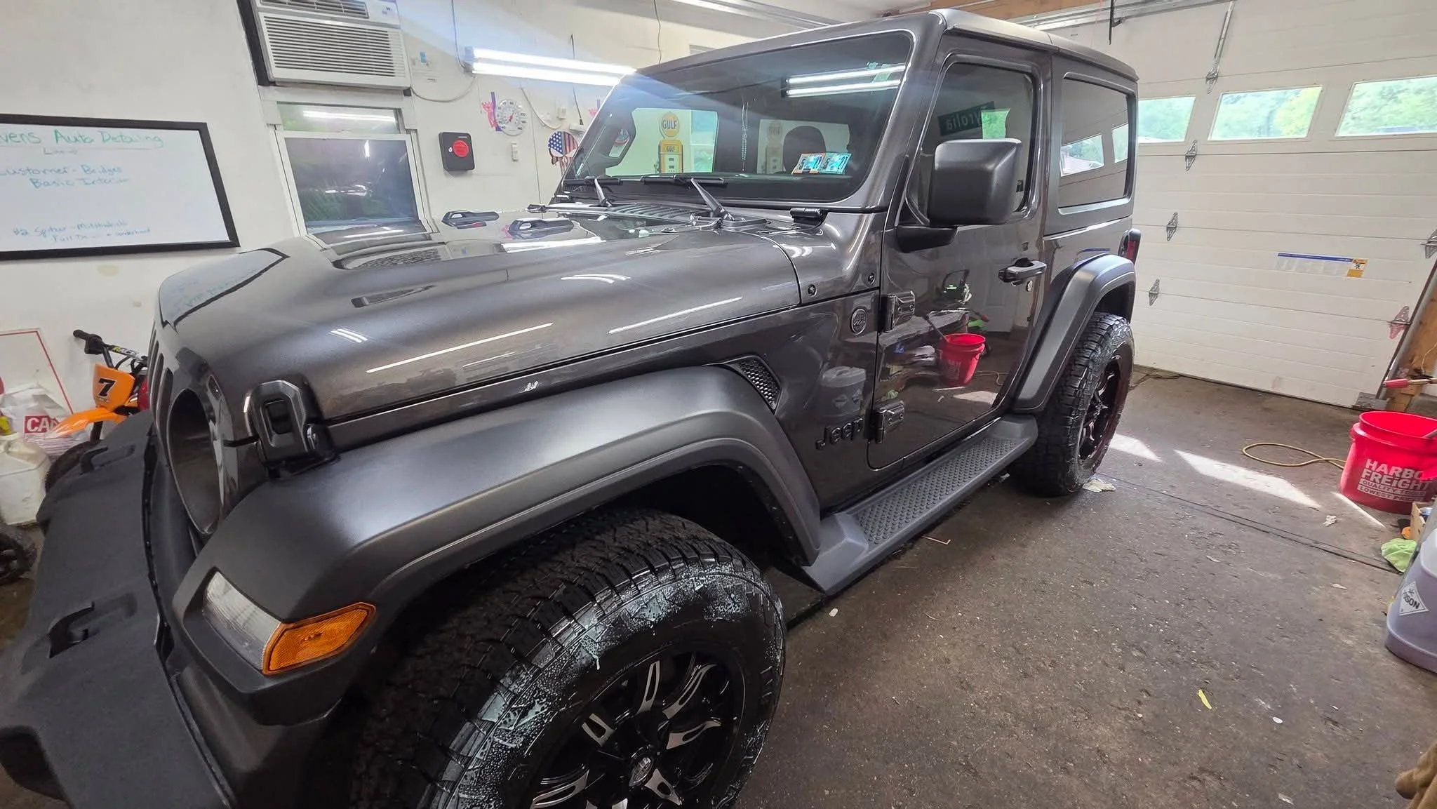 A gray Jeep in a garage with a white garage door, various tools, a red bucket, and windows in the background.