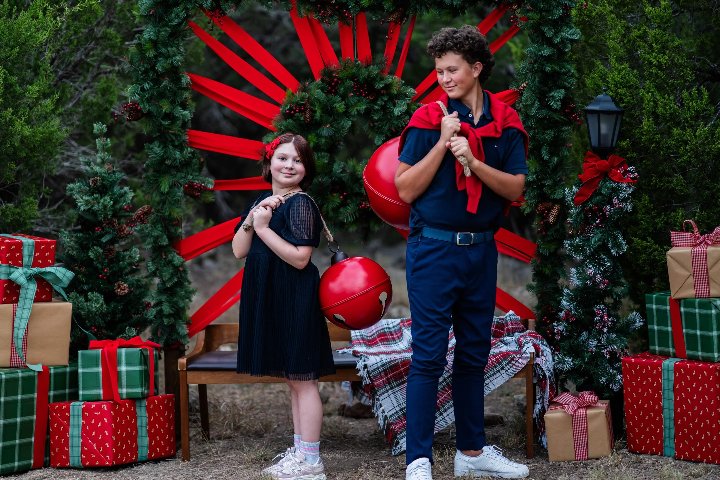 Two children, a girl and a boy, standing outdoors in front of a Christmas wreath decorated with red ribbons and ornaments, surrounded by wrapped presents, with a small bench and a plaid blanket on the ground.