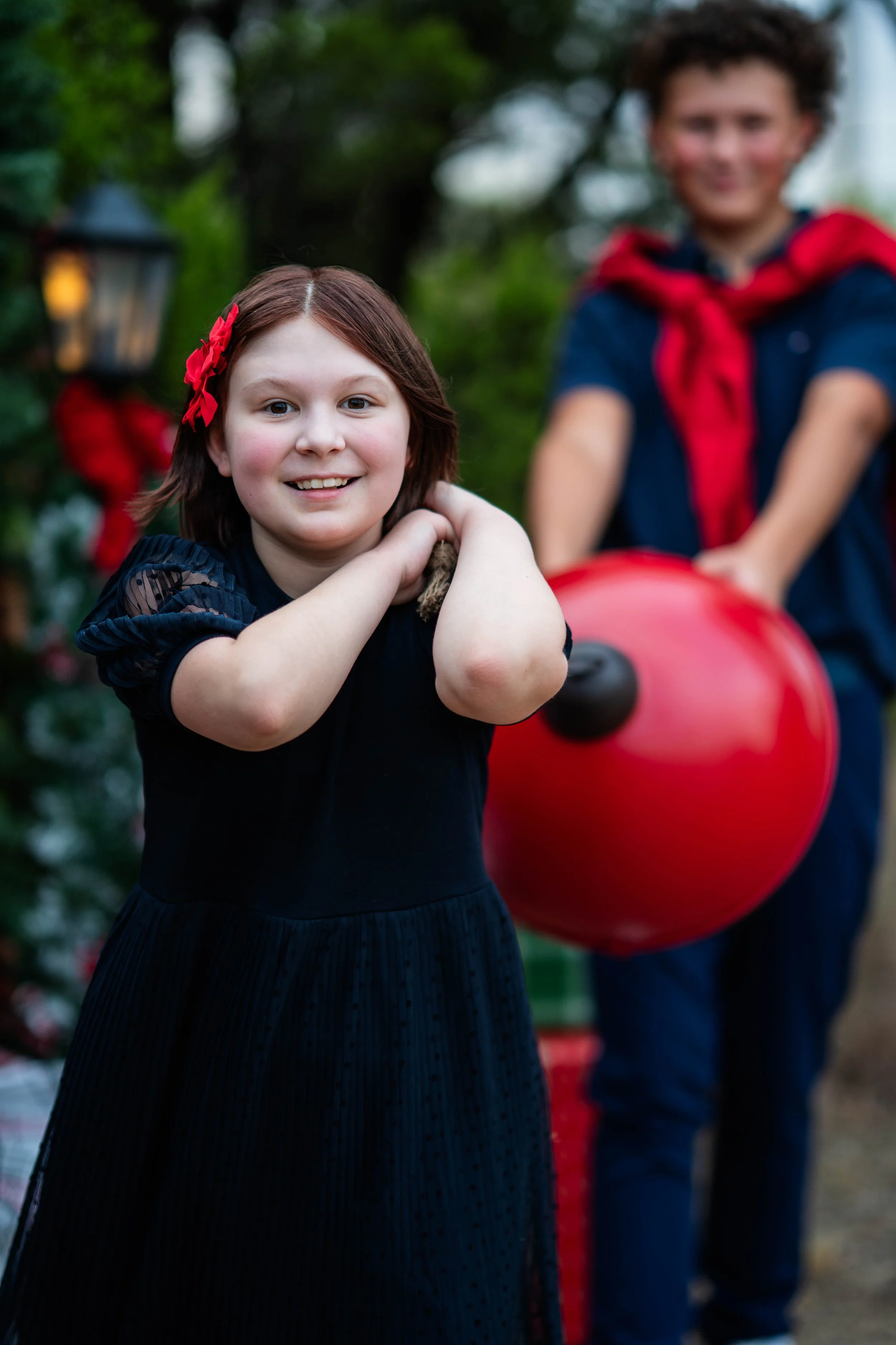 A young girl smiling and holding her arms crossed near her shoulders, standing outdoors with a boy behind her holding a large red balloon. The girl has shoulder-length brown hair with a red bow, and both are dressed casually. The background is blurred with greenery and outdoor decorations.