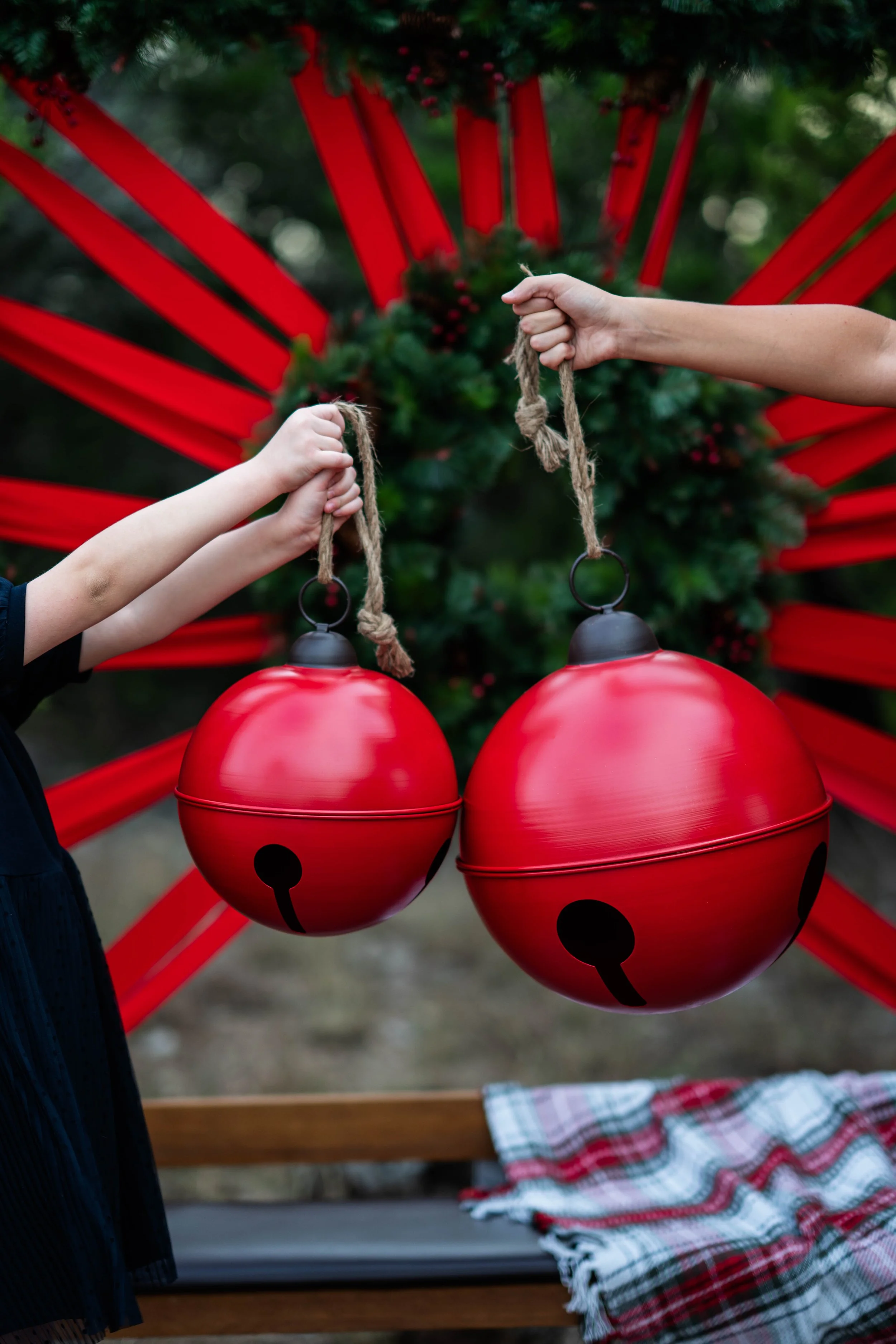 Two people hold red Christmas ornaments shaped like jingle bells in front of a decorated Christmas tree with a red backdrop.