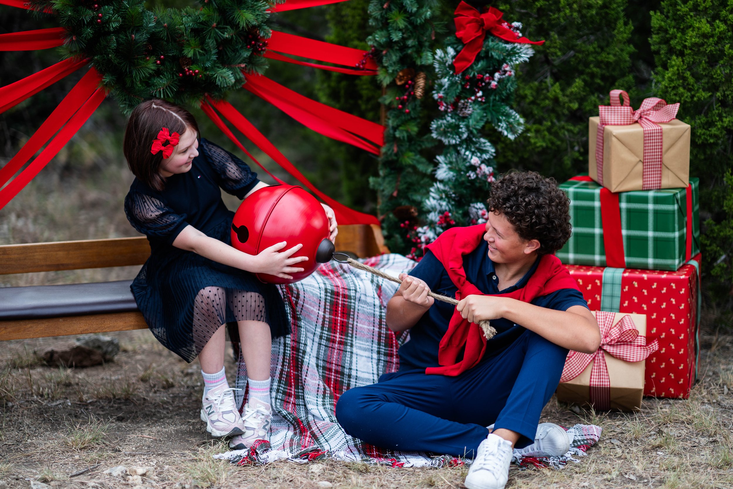 A girl and a boy playing tug of war with a red toy bomb outdoors during Christmas, with decorated Christmas trees and wrapped presents behind them.