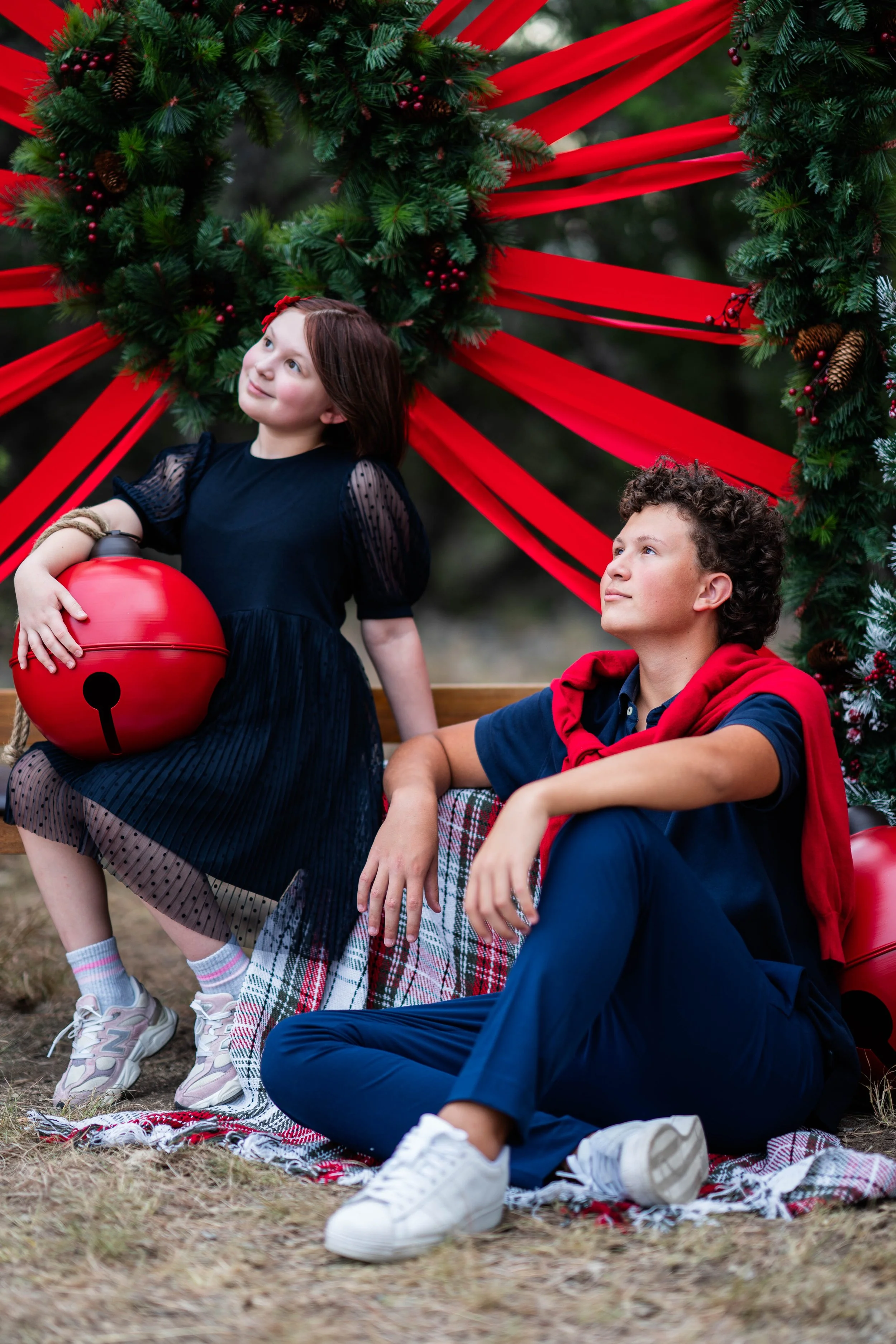 A young girl holding a red Christmas ornament bucket and a boy sitting on a blanket with Christmas decorations in the background.