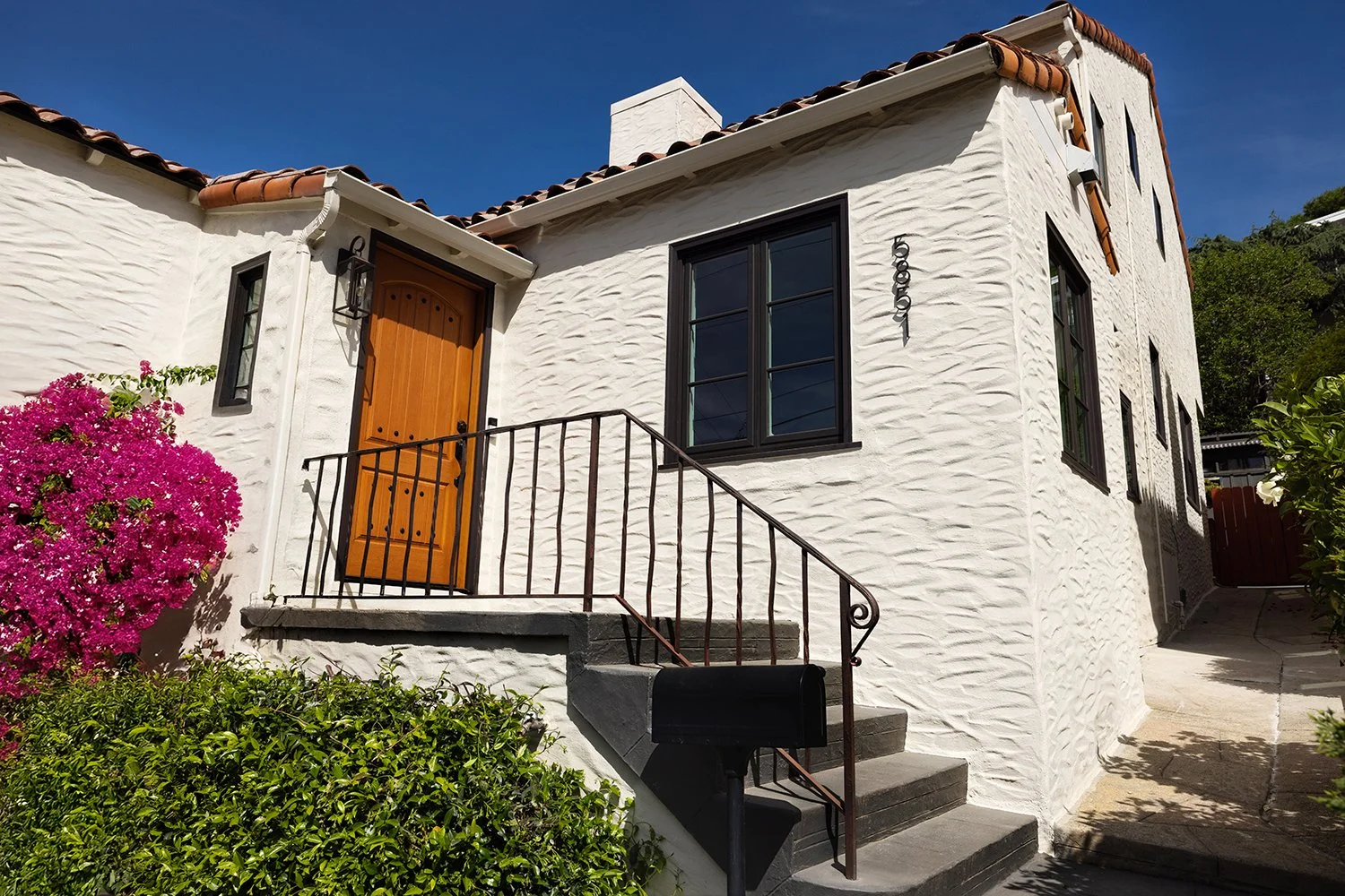 White stucco house with a wooden front door, black-framed windows, a small staircase with a black railing, a pink flowering bush, and greenery under clear blue sky.