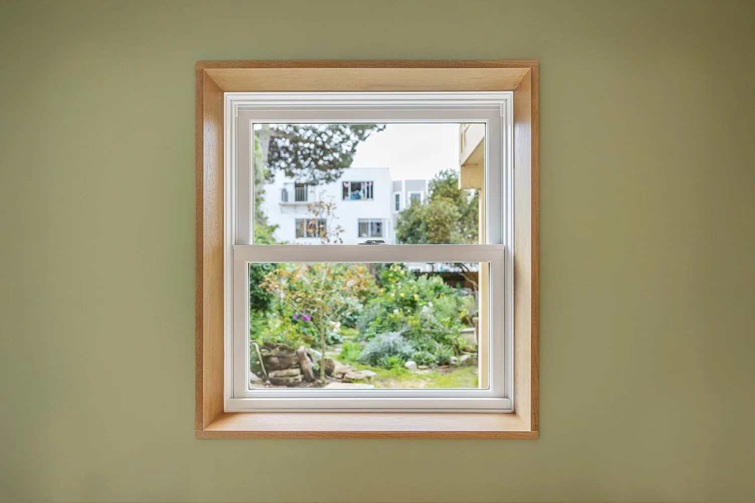 View of a garden and modern white apartment building through a small double-pane window on a green wall.