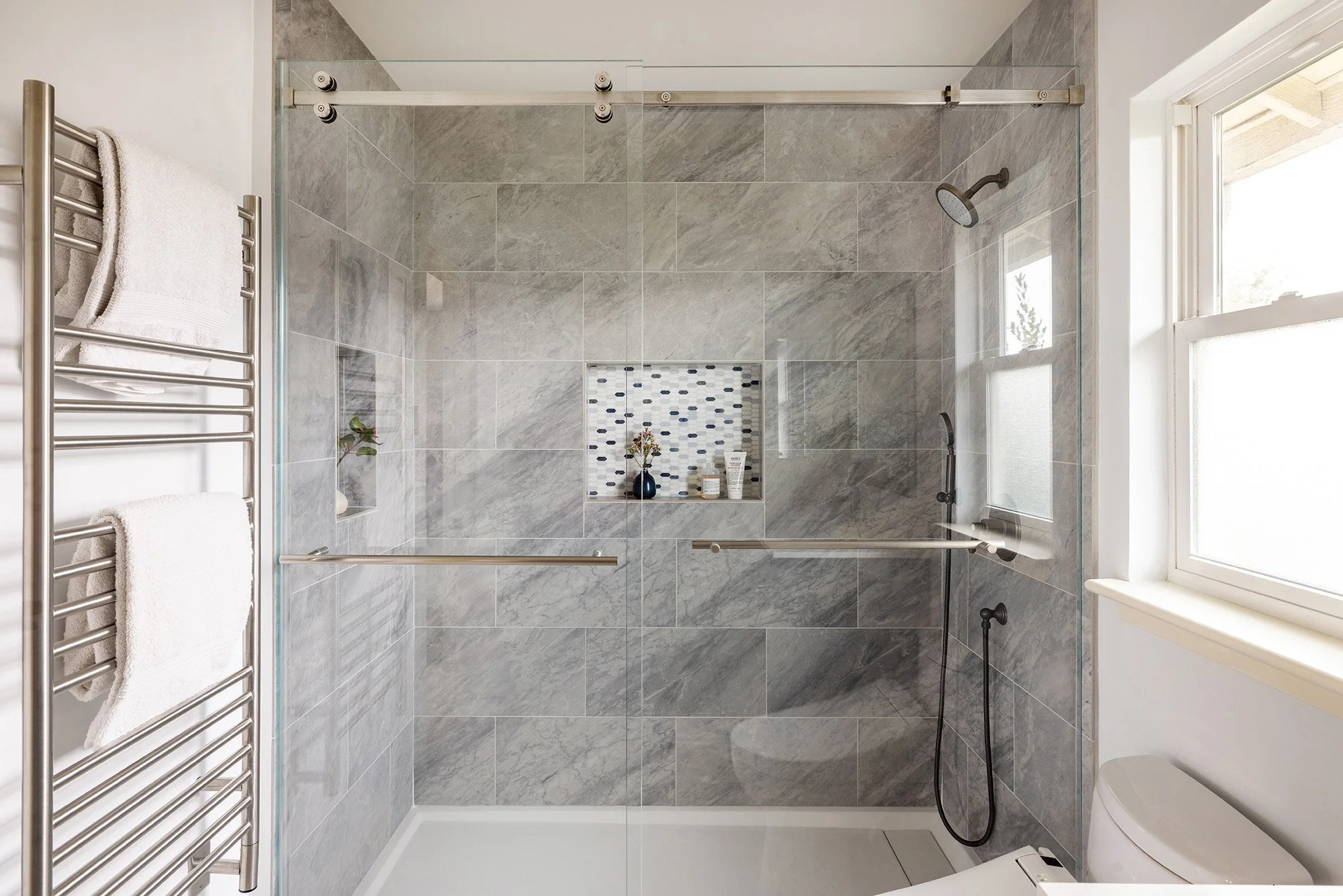 Modern walk-in shower with gray tile walls, glass door, built-in shelves, and showerheads, next to a window.