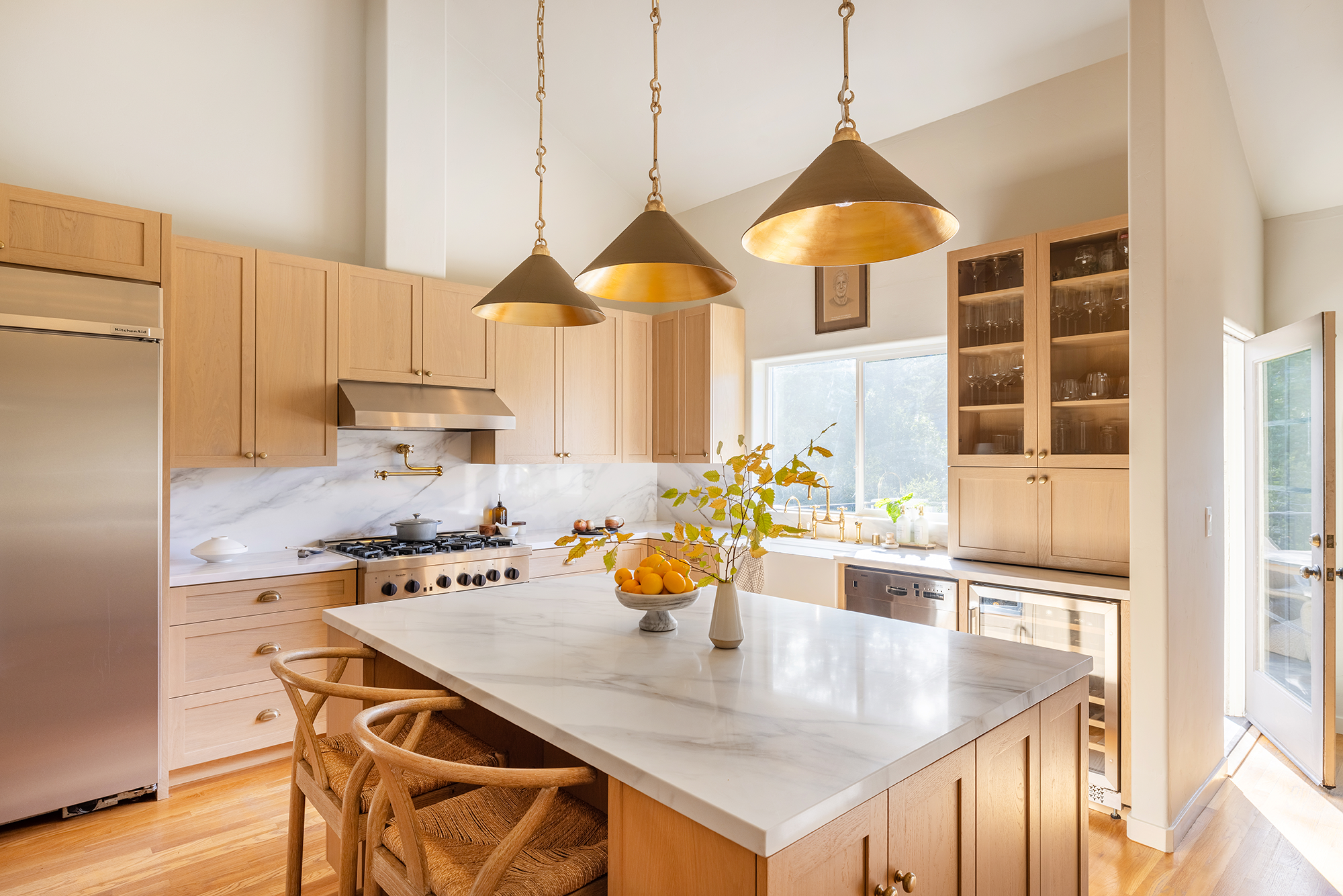A modern kitchen with light wood cabinets, a white marble countertop, and a white marble backsplash. There are three gold pendant lights hanging above the center island. The island has a white marble top with a bowl of lemons and a white vase with green leafy branches. To the right, there are glass-front cabinets with glassware, and a window letting in natural light. A stainless steel refrigerator is partially visible on the left.