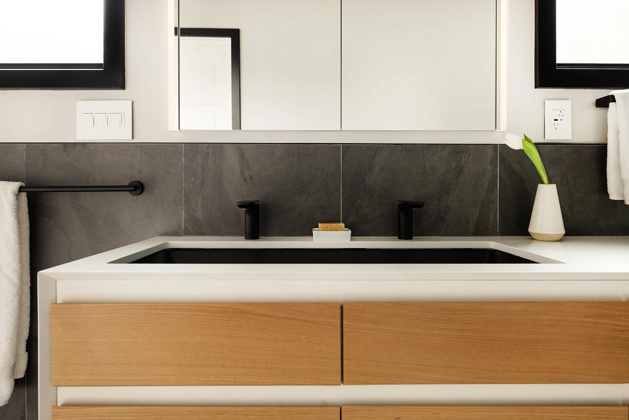A modern bathroom vanity with a white countertop, black faucets, a large mirror, and a small potted plant with a white flower on a beige vase.