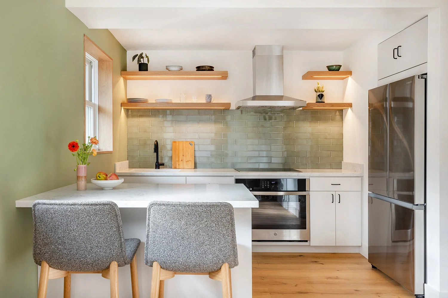 Modern kitchen with gray and white cabinetry, green tile backsplash, open wooden shelves, stainless steel appliances, and a small dining area with two gray chairs.