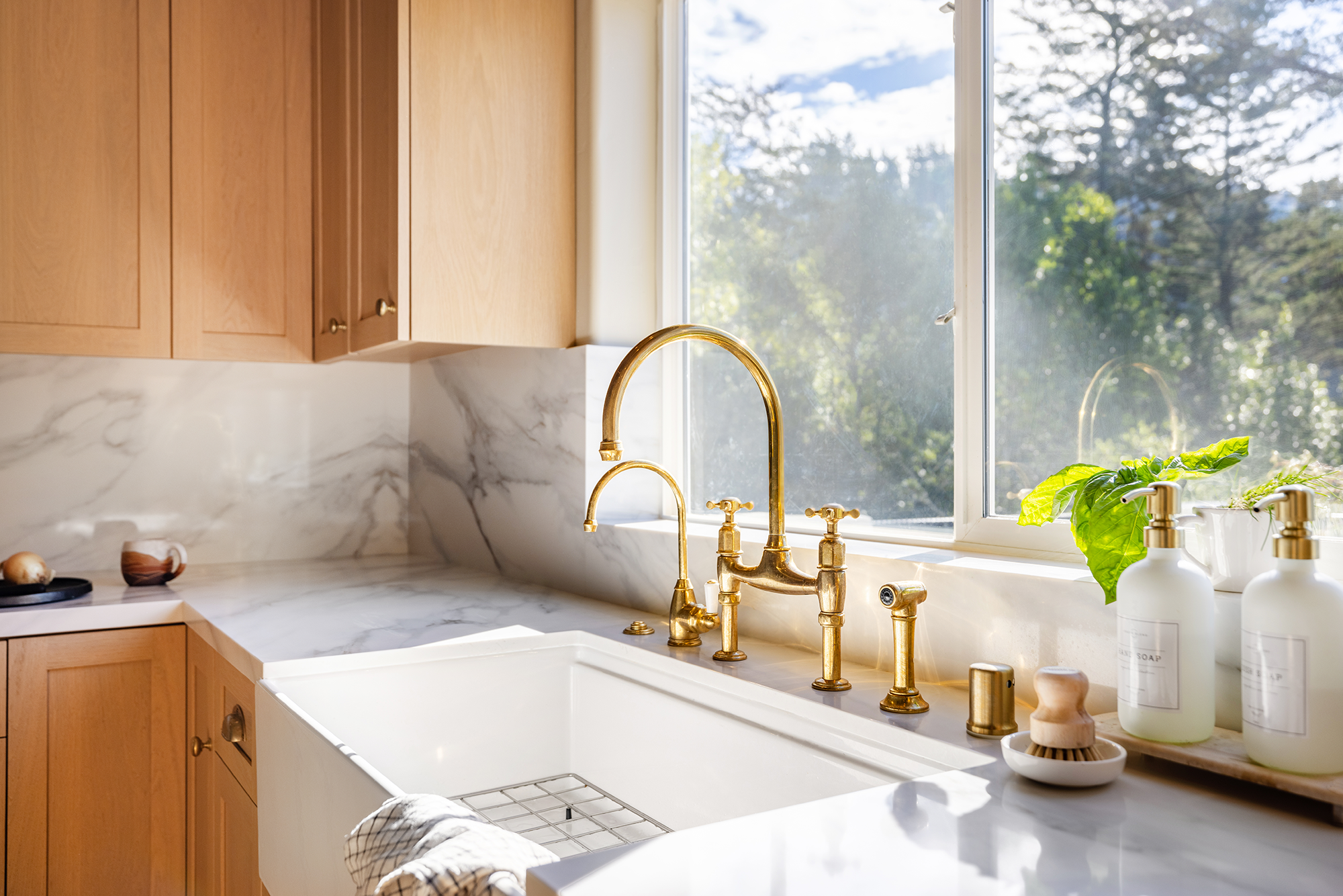 A bright kitchen with wooden cabinets, a white marble countertop, and a large window with a view of trees outside. There is a gold faucet over a white sink, and various soap dispensers and a plant on the counter.