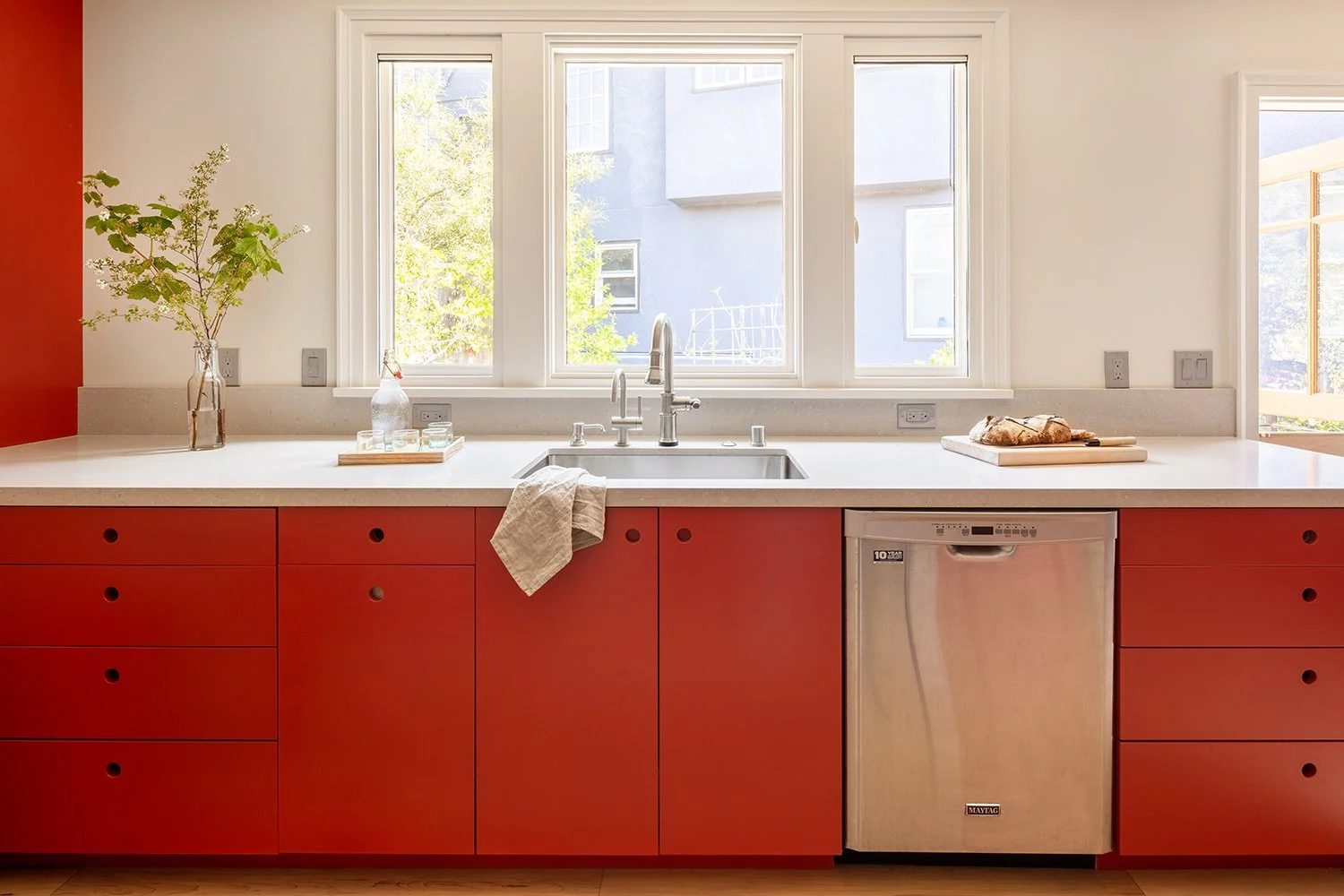 Modern kitchen with red cabinets, a white countertop, and a stainless steel dishwasher. A window above the sink lets in natural light, with a vase of green branches and a tray with glass bottles and a glass on the counter. A towel hangs on the front of one cabinet, and a cutting board with bread is on the right side of the counter.