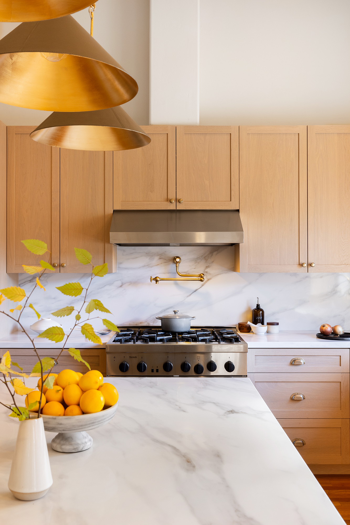 A modern kitchen with light wood cabinets, a stainless steel stove, and a marble backsplash. There is a bowl of lemons on the white marble countertop in the foreground, and some kitchen accessories on the countertop.
