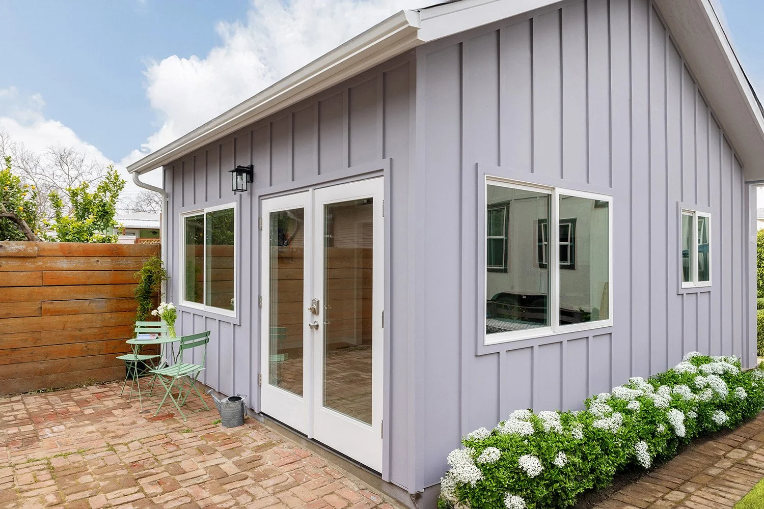 Small, light purple shed with white-framed windows and a glass door, situated on a brick patio with white flowers along its base and a wooden fence with a tree behind it, under a partly cloudy sky.