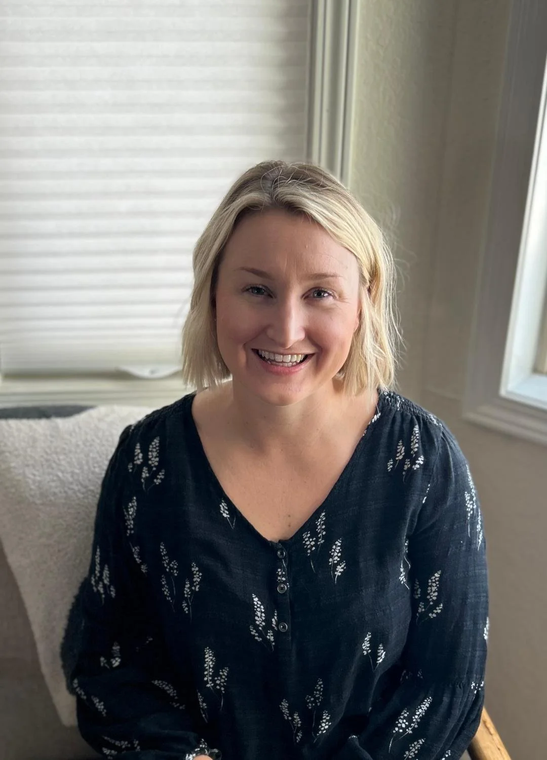 A woman with short blonde hair is smiling and sitting on a chair near a window with closed blinds.