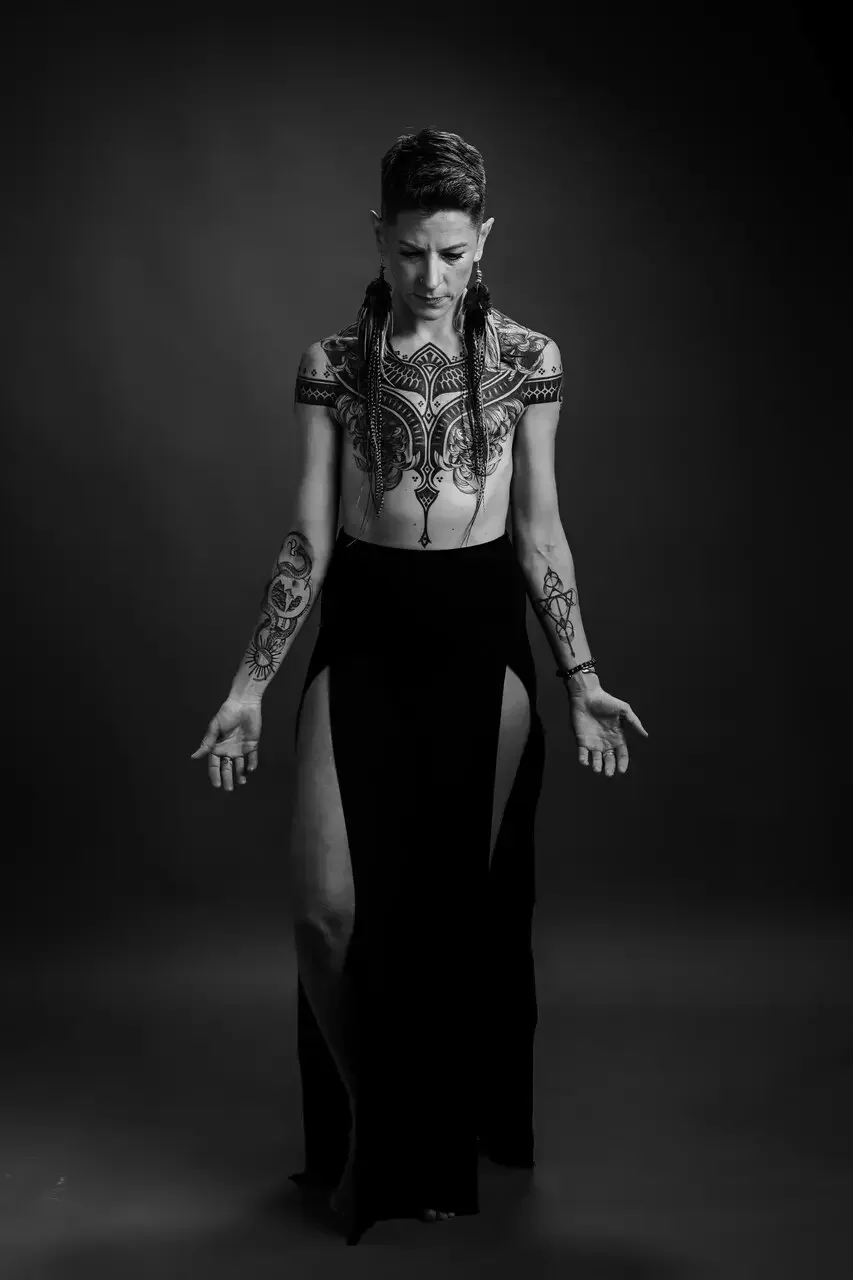 A black and white portrait of Dr. Kallie Harrison showcasing her full chest and shoulder tattoos while standing in a meditative pose.