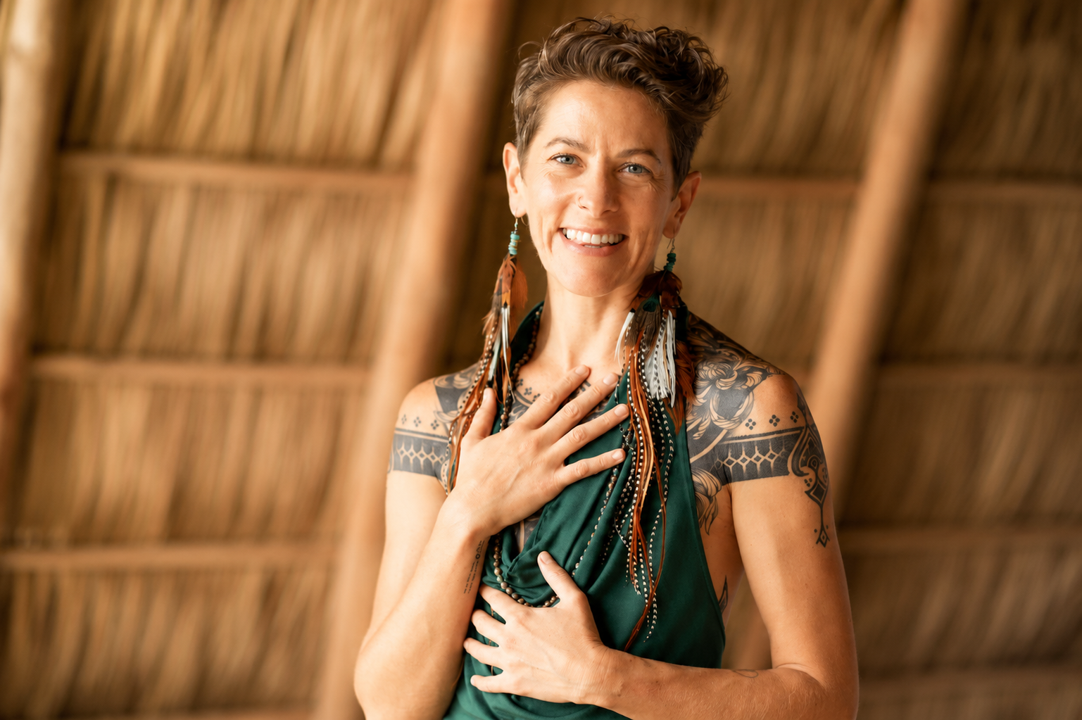 A smiling woman with short curly hair and tattoos on her arms and shoulders, wearing feather earrings and a green dress, standing inside a thatched-roof structure.
